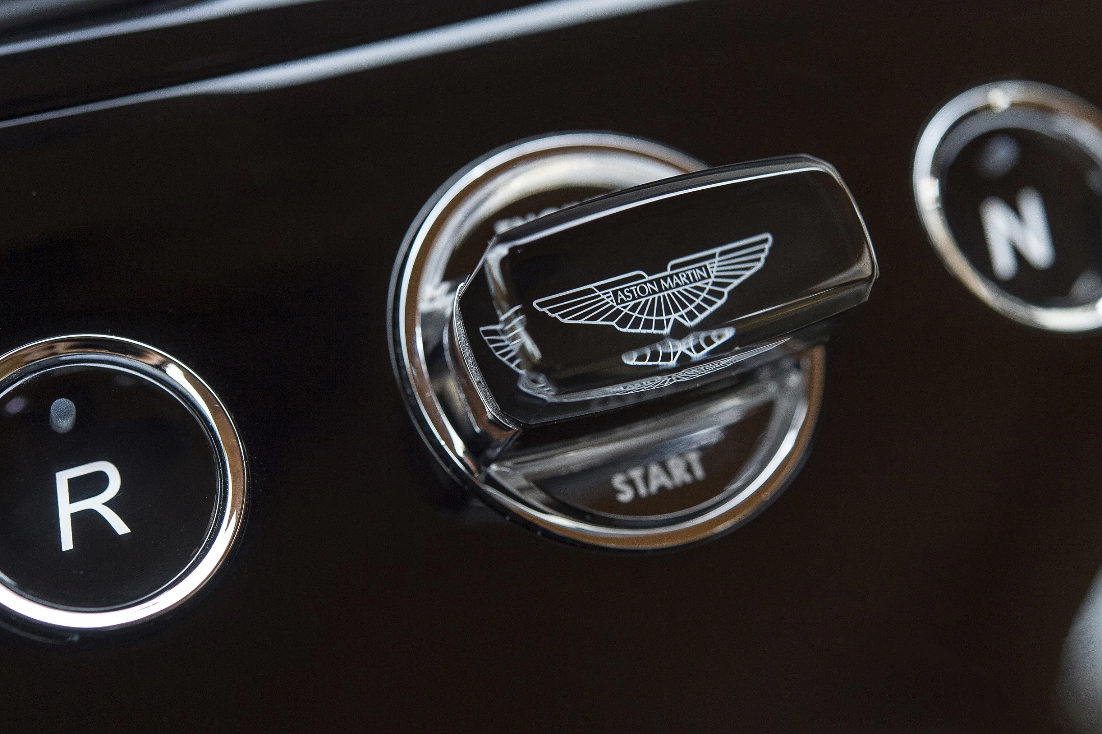 Nahaufnahme eines Zündschalters eines Aston Martin mit markantem Flügel-Emblem auf schwarzem Armaturenbrett.