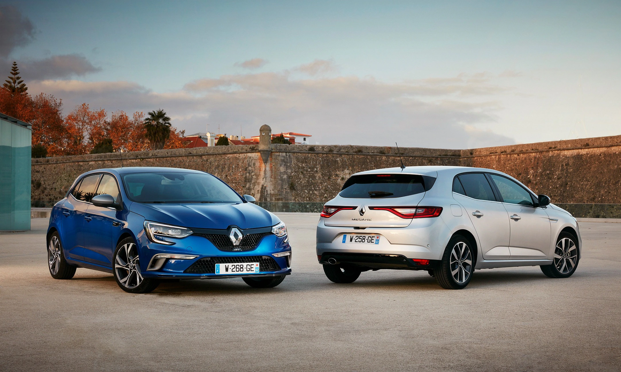 Zwei Renault Megane Autos, blau und weiß, parken im Freien vor einer Steinmauer bei Sonnenuntergang.