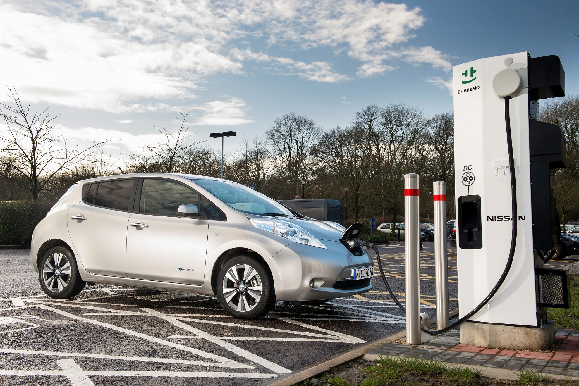 Silbernes Elektroauto lädt an öffentlicher Ladestation auf einem Parkplatz bei bewölktem Himmel und Bäumen im Hintergrund.