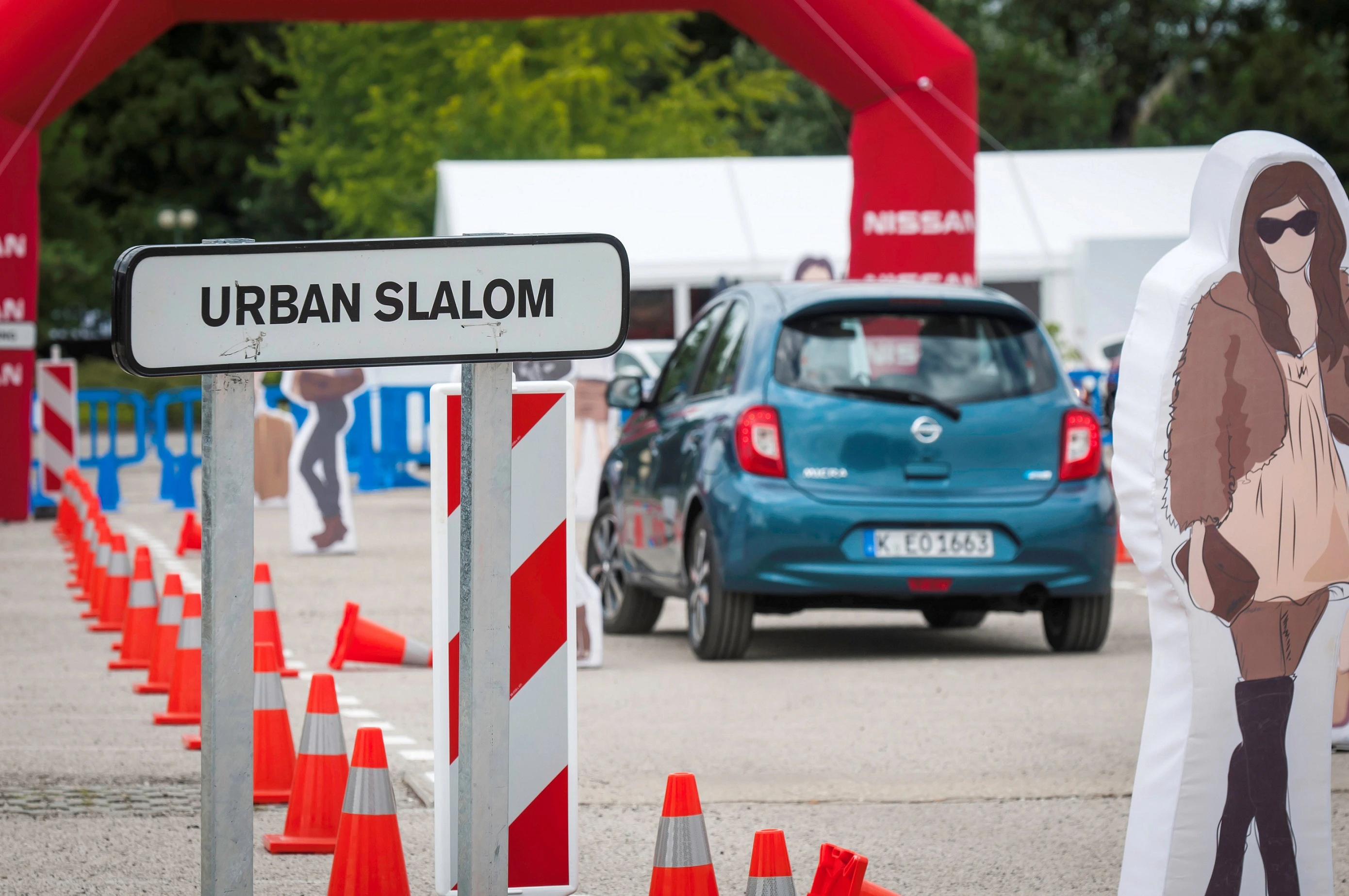 Ein Auto fährt durch einen Urban Slalom Parcours mit Verkehrskegeln und Pappfiguren. Ein Schild steht im Vordergrund.