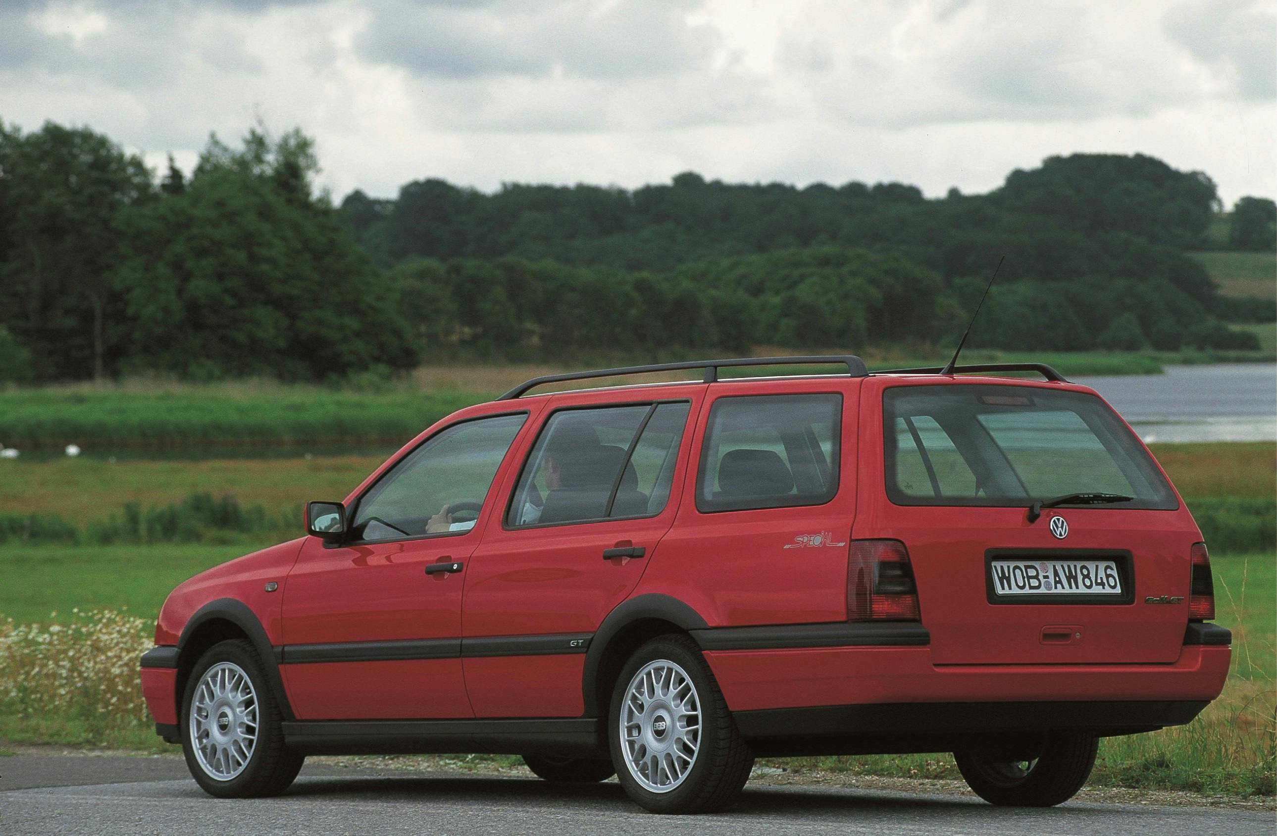 Roter Kombi auf Landstraße, umgeben von Wiesen und einem See im Hintergrund, unter bewölktem Himmel.