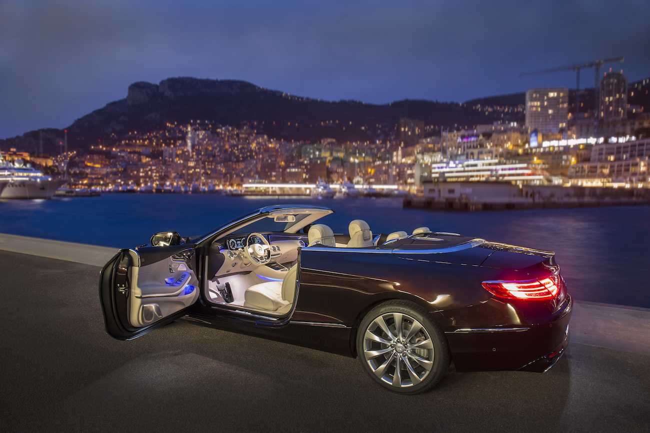 Luxuriöses Cabriolet mit offener Tür, parkend an einer Küste bei Nacht. Lichter der Stadt und Yachten im Hintergrund.