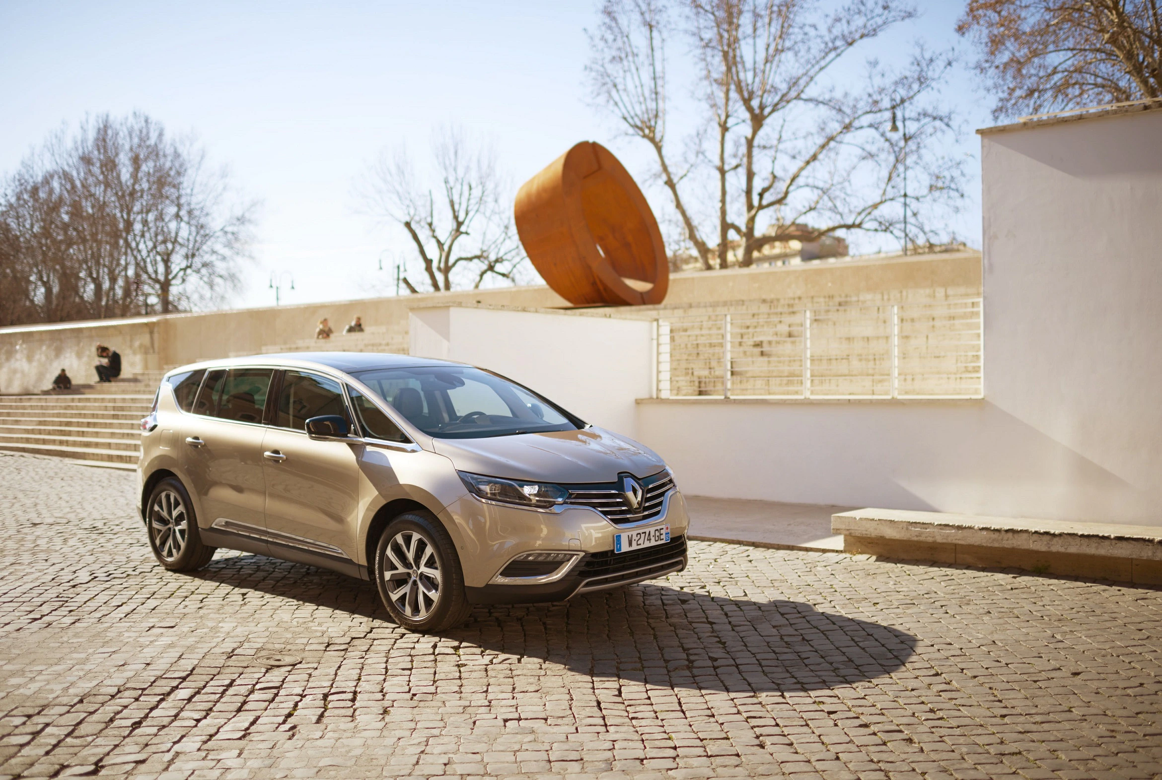 Goldener Minivan steht auf gepflasterter Straße bei sonnigem Wetter, mit kahlen Bäumen und moderner Skulptur im Hintergrund.