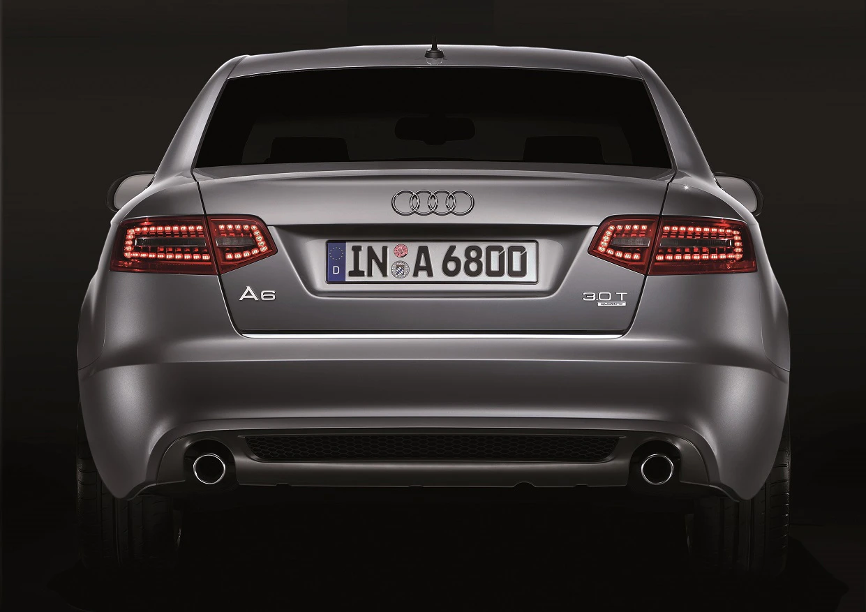 Rückansicht eines grauen Audi A6 mit deutschen Kennzeichen auf schwarzem Hintergrund. LED-Rückleuchten und Doppelrohrauspuff sind sichtbar.