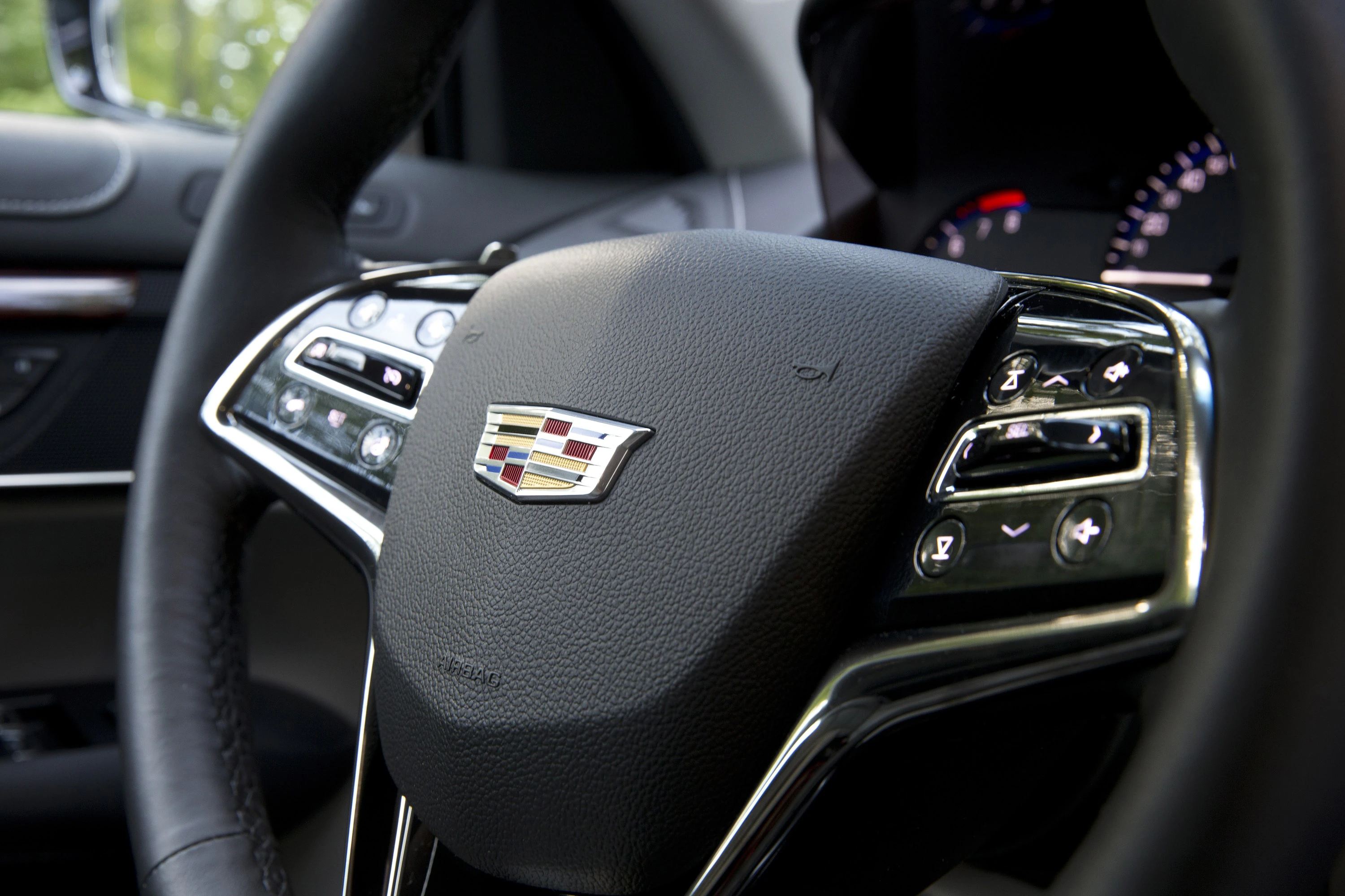 Nahaufnahme eines Cadillac-Lenkrads mit eingebauten Bedienelementen und Markenlogo in der Mitte. Instrumententafel im Hintergrund.