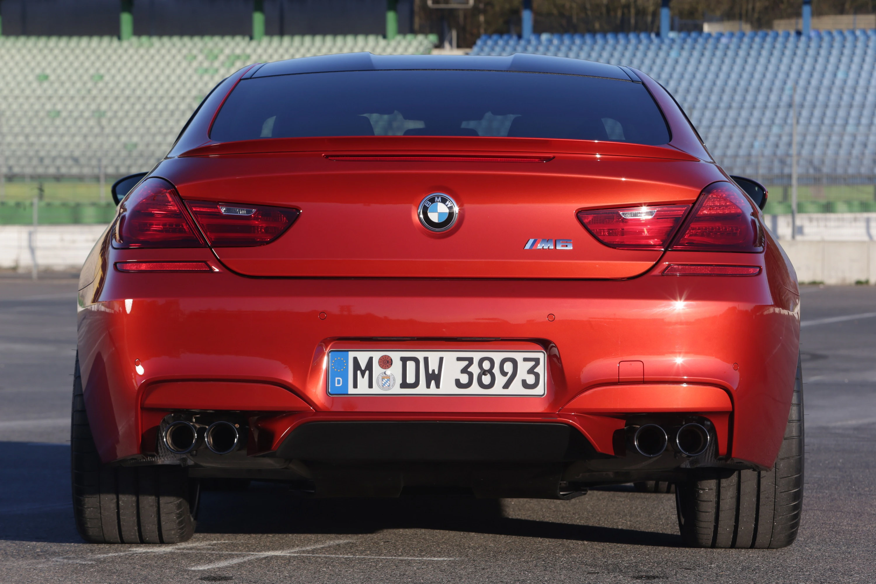 Roter BMW M6 von hinten, geparkt auf freier Fläche, mit erkennbaren Rücklichtern und Auspuffen. Hintergrund: leere Tribünen.