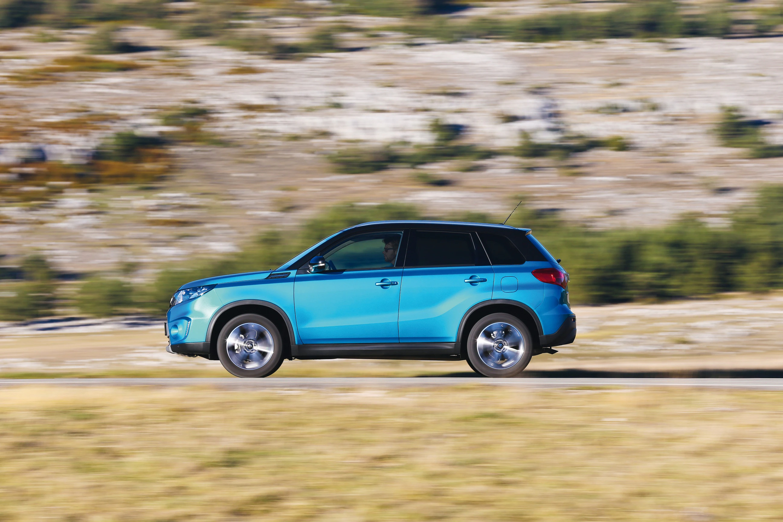 Blauer SUV fährt schnell auf einer Landstraße durch eine hügelige Landschaft mit verschwommenem Hintergrund aus Felsen und Bäumen.