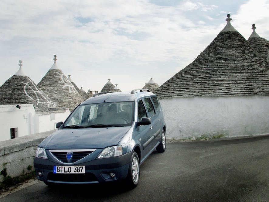 Blauer Kombi vor traditionellen Trulli-Häusern mit kegelförmigen Dächern, unter bewölktem Himmel.