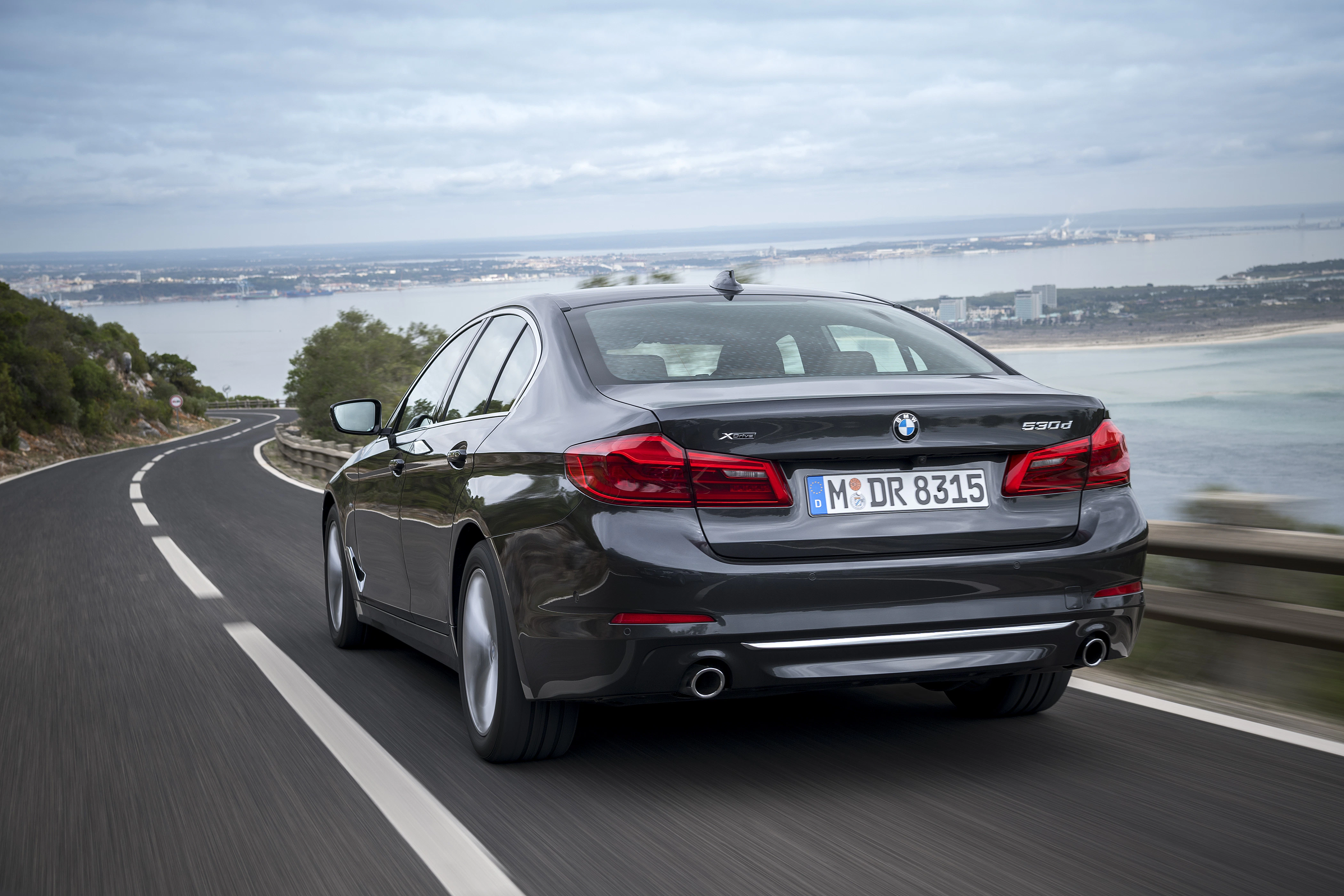BMW-Limousine fährt auf einer kurvigen Küstenstraße mit Meerblick im Hintergrund. Bewölkter Himmel und Natur entlang der Straße.