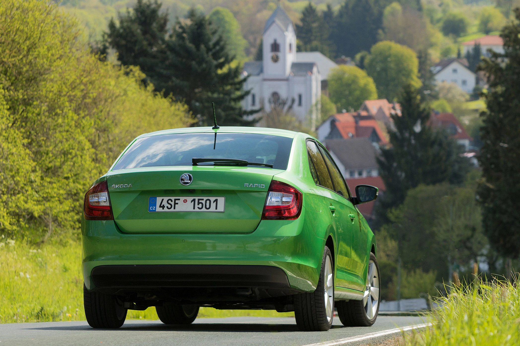 Grünes Auto auf Landstraße mit Blick auf Dorf und Kirche im Hintergrund, umgeben von Bäumen und hügeliger Landschaft.
