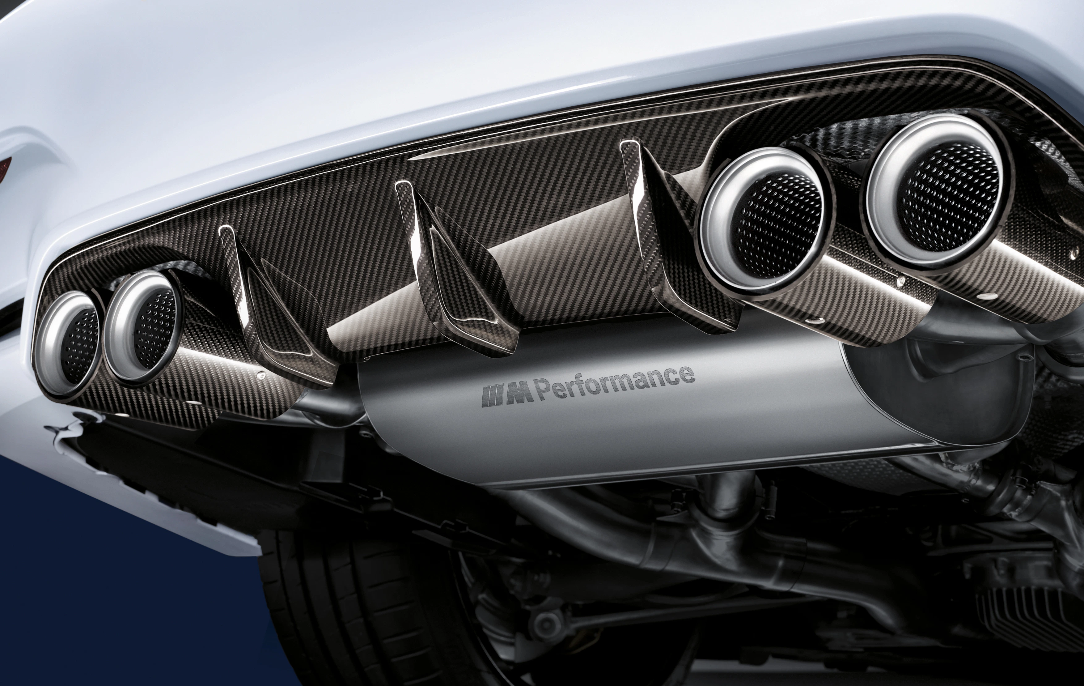 Nahaufnahme eines sportlichen Autoauspuffs mit vier verchromten Endrohren und Carbon-Details vor blauem Hintergrund.