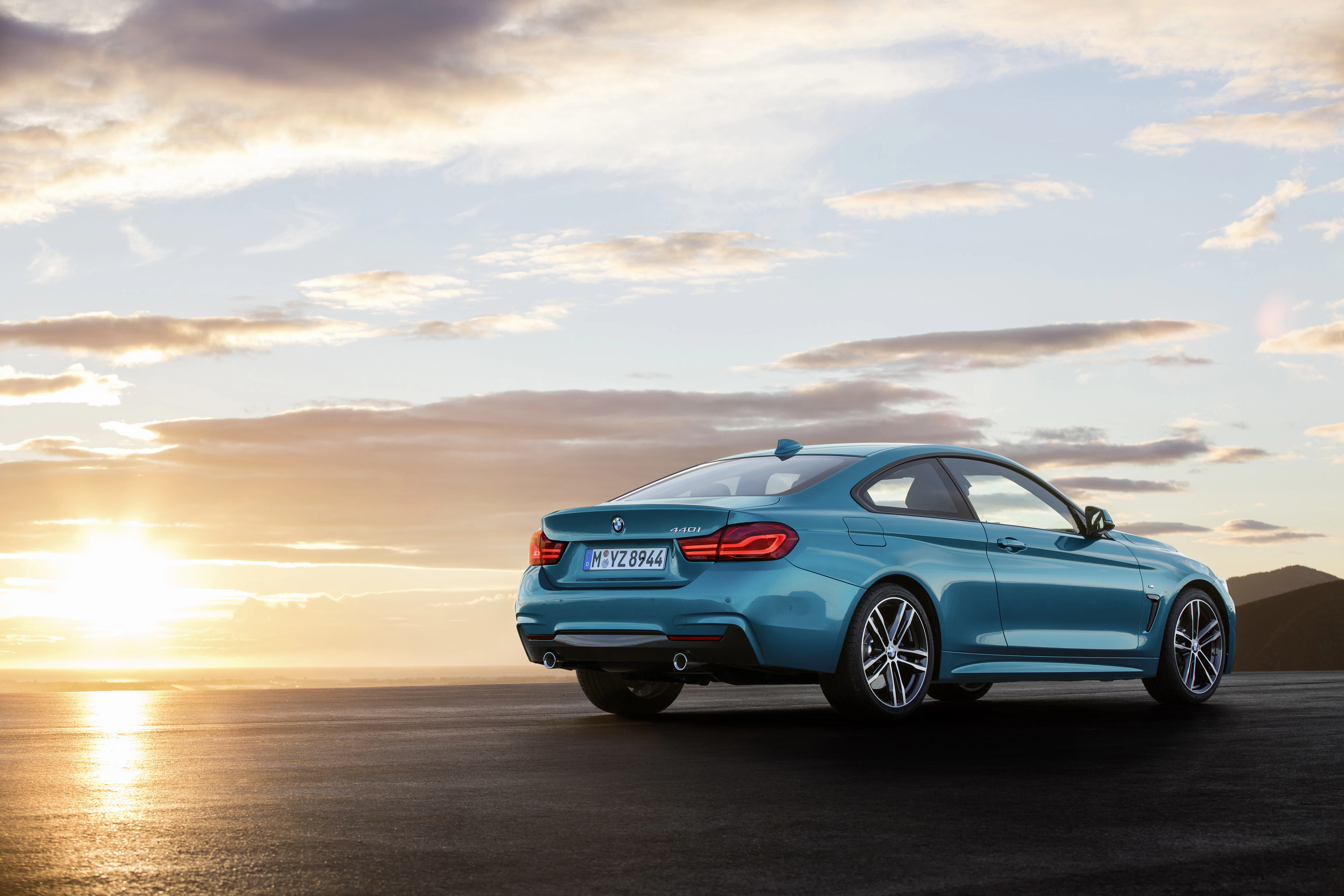 Blaues Sportcoupé auf offener Straße bei Sonnenuntergang, leichte Wolken am Himmel, Asphalt im Vordergrund.