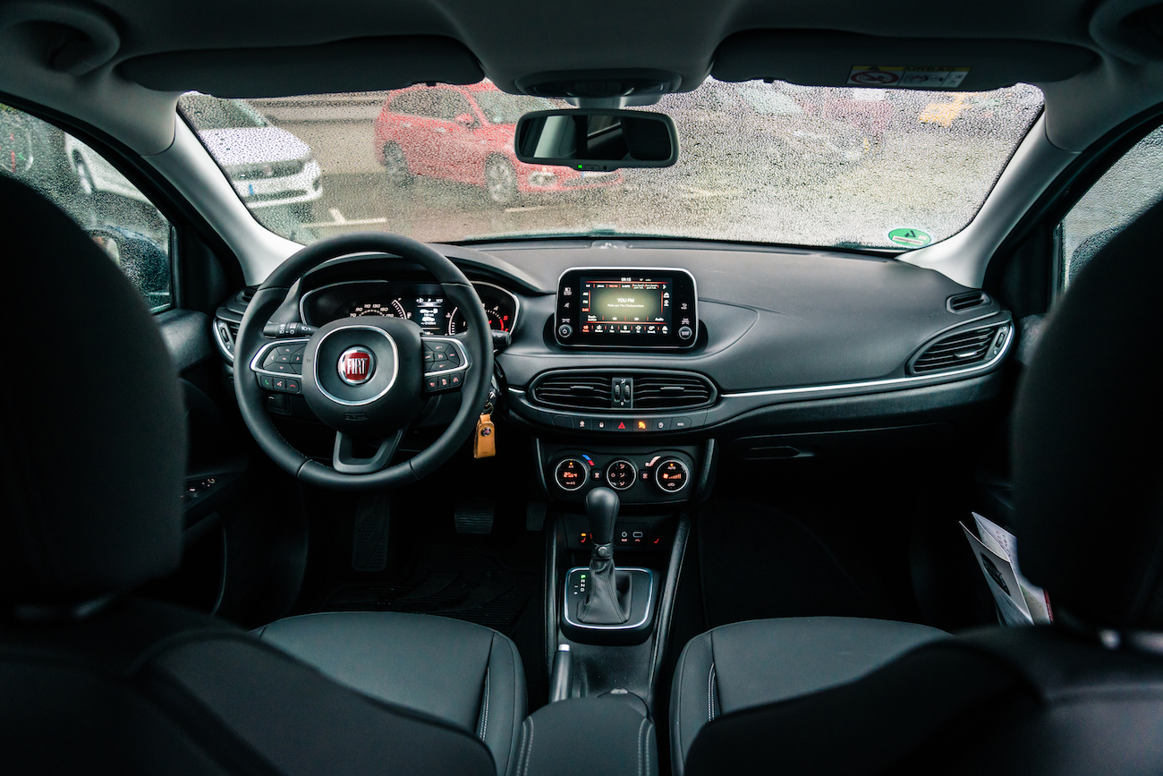 Innenraum eines Autos mit modernen Armaturen, Lenkrad links und Touchscreen-Display in der Mittelkonsole. Regentropfen auf der Windschutzscheibe.