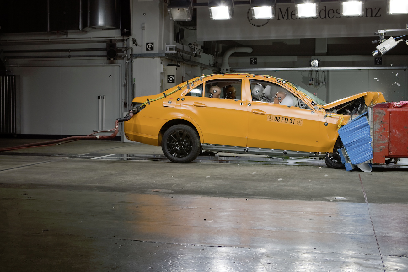 Gelbes Auto bei Crashtest in einer Halle, Frontpartie stark deformiert, Dummies im Innenraum, blaue Schutzpolster vorne.