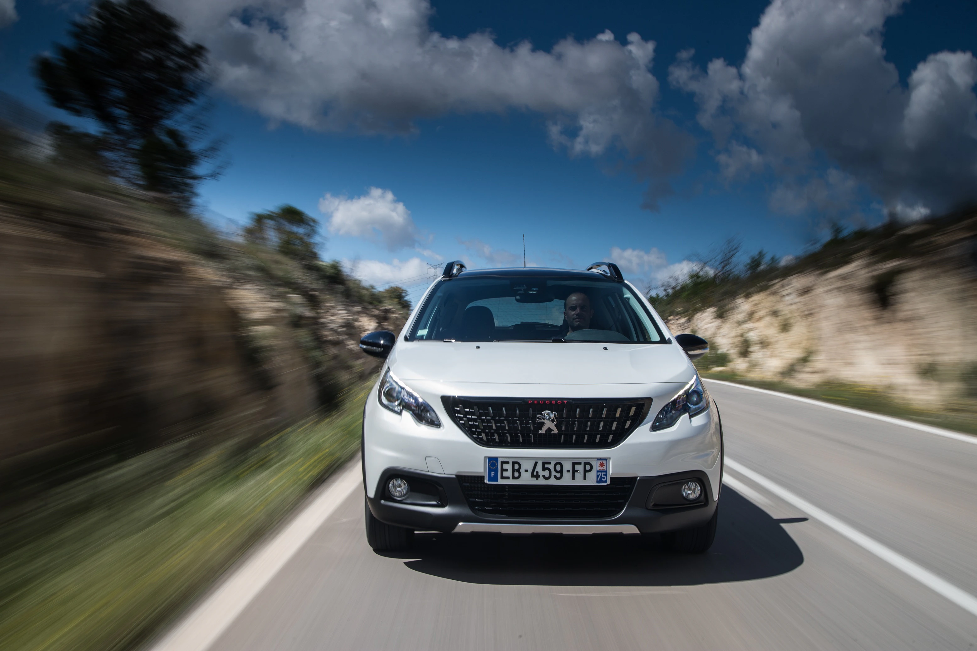 Weißer Peugeot fährt auf einsamer, kurviger Landstraße bei bewölktem Himmel. Dynamische Perspektive von vorne.