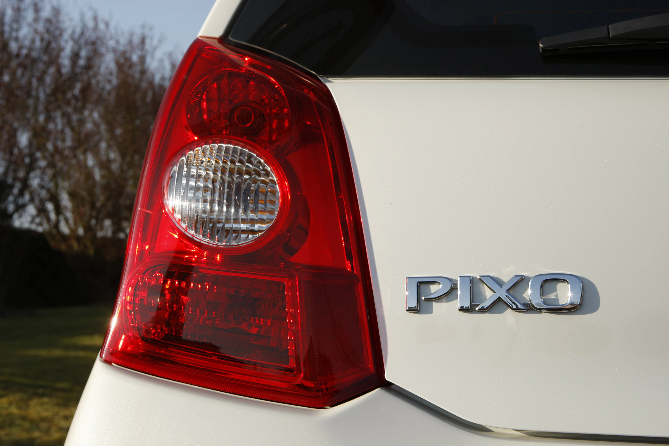 Nahaufnahme eines weißen Autos mit rotem Rücklicht und dem Schriftzug "PIXO" am Heck. Hintergrund unscharf mit Bäumen.