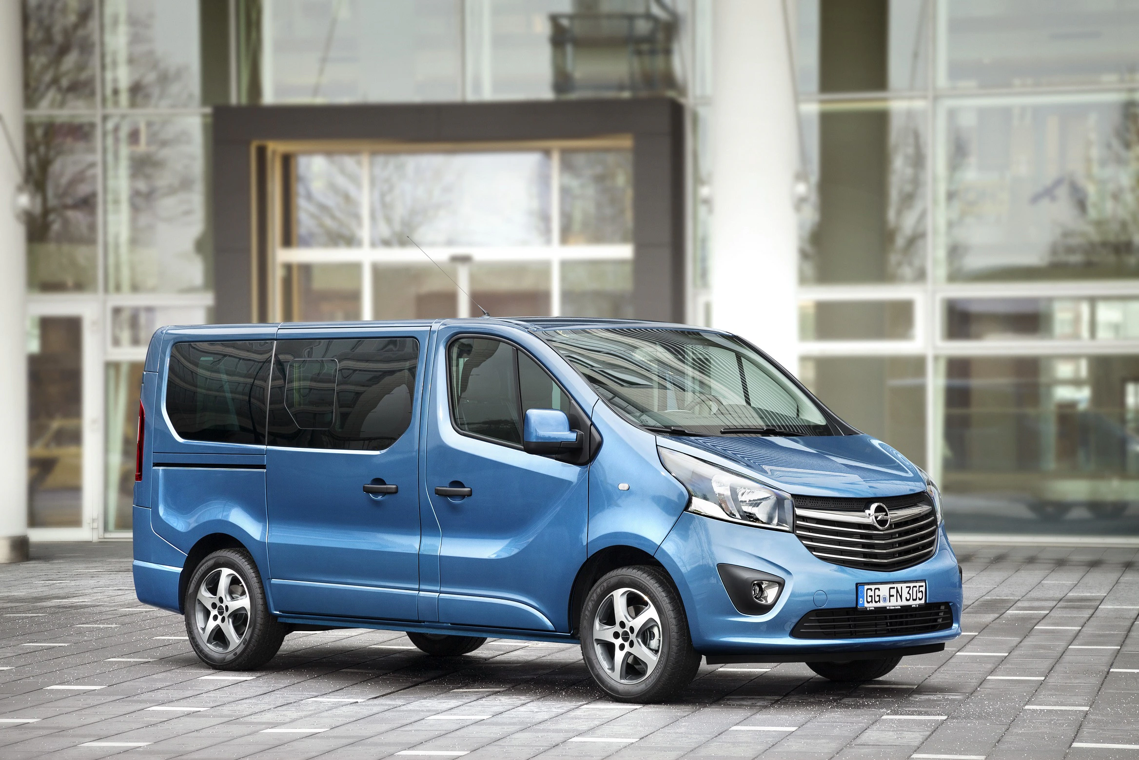 Blauer Minivan, geparkt auf einem gepflasterten Platz vor einem modernen Gebäude mit großen Glasfenstern.
