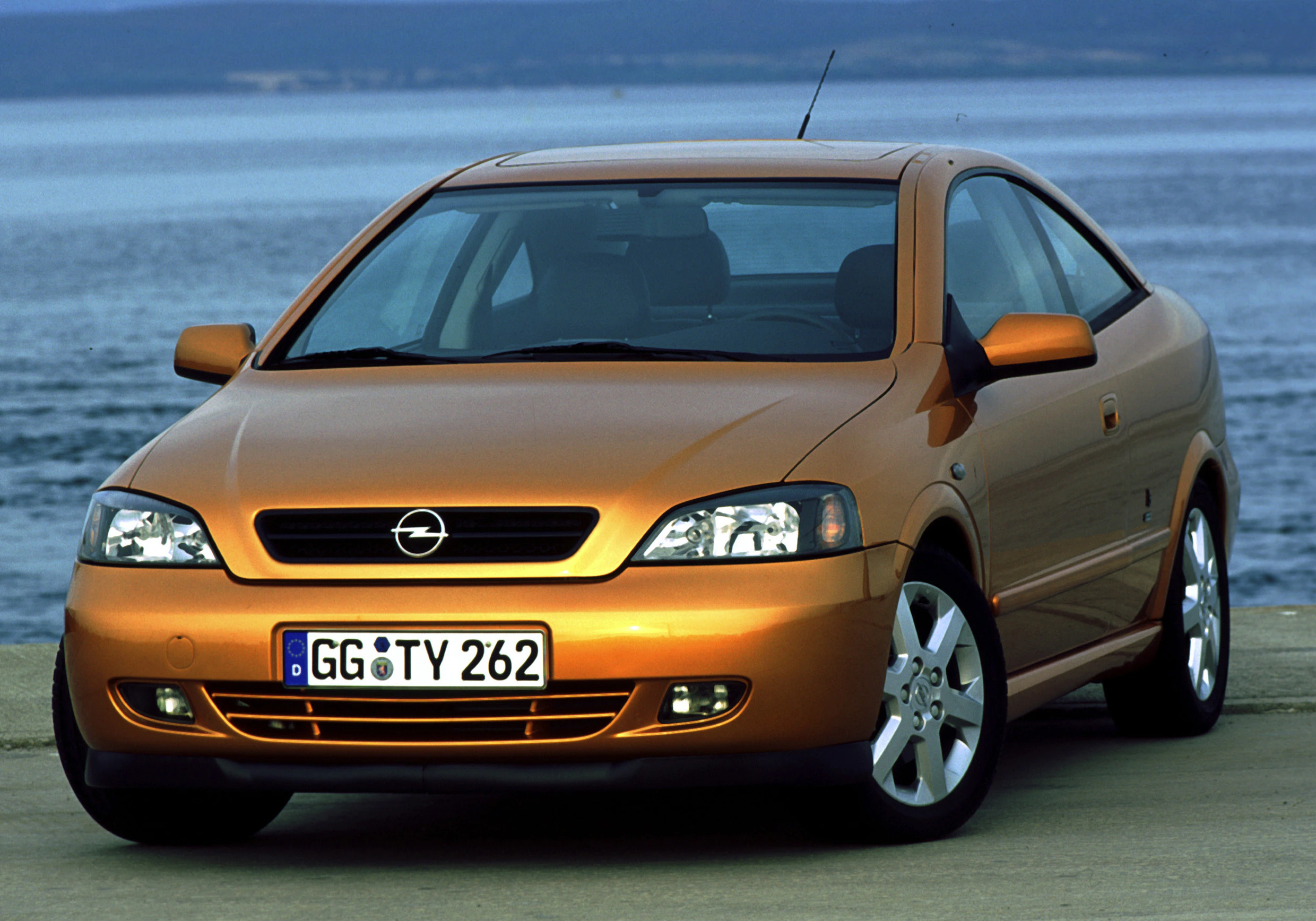 Orangefarbener Opel-Coupé parkt vor einem See oder Meer. Kühlergrill mit Logo, Scheinwerfer eingeschaltet, klare Himmelsspiegelung auf Wasser.