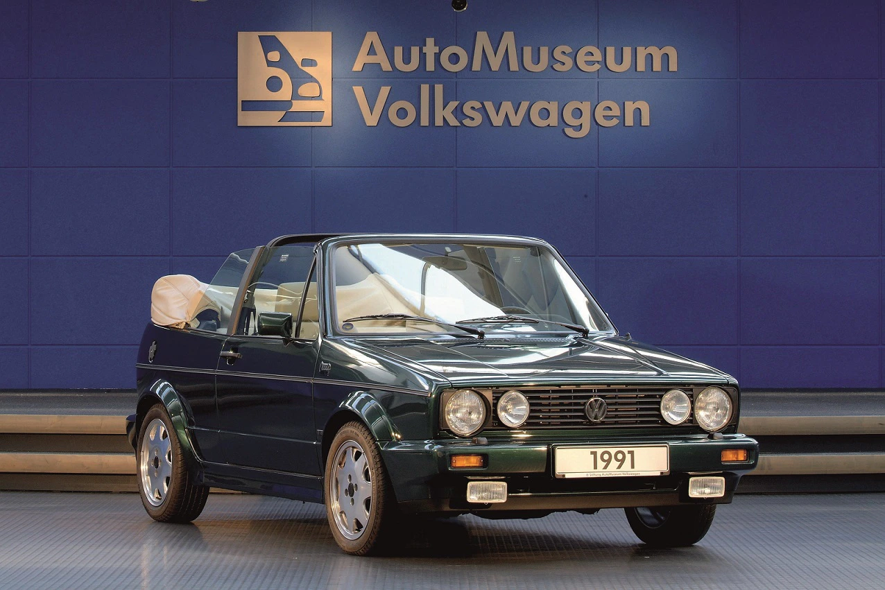 Ein dunkelgrüner VW Cabrio von 1991 im AutoMuseum Volkswagen, frontal fotografiert, auf einer Ausstellungsbühne mit blauem Hintergrund.