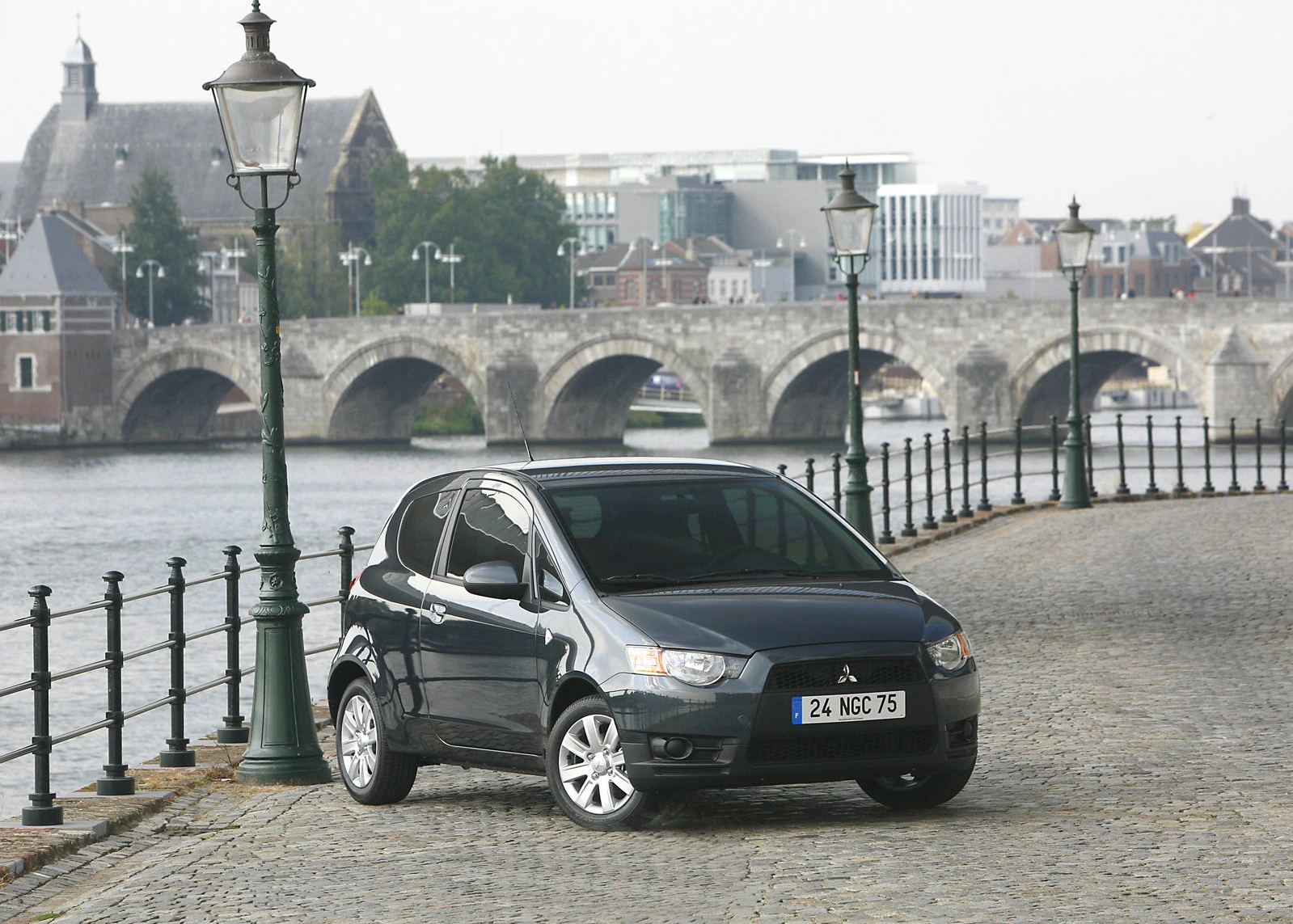 Schwarzes Auto parkt auf Kopfsteinpflaster am Fluss, im Hintergrund eine historische Steinbrücke und alte Gebäude.