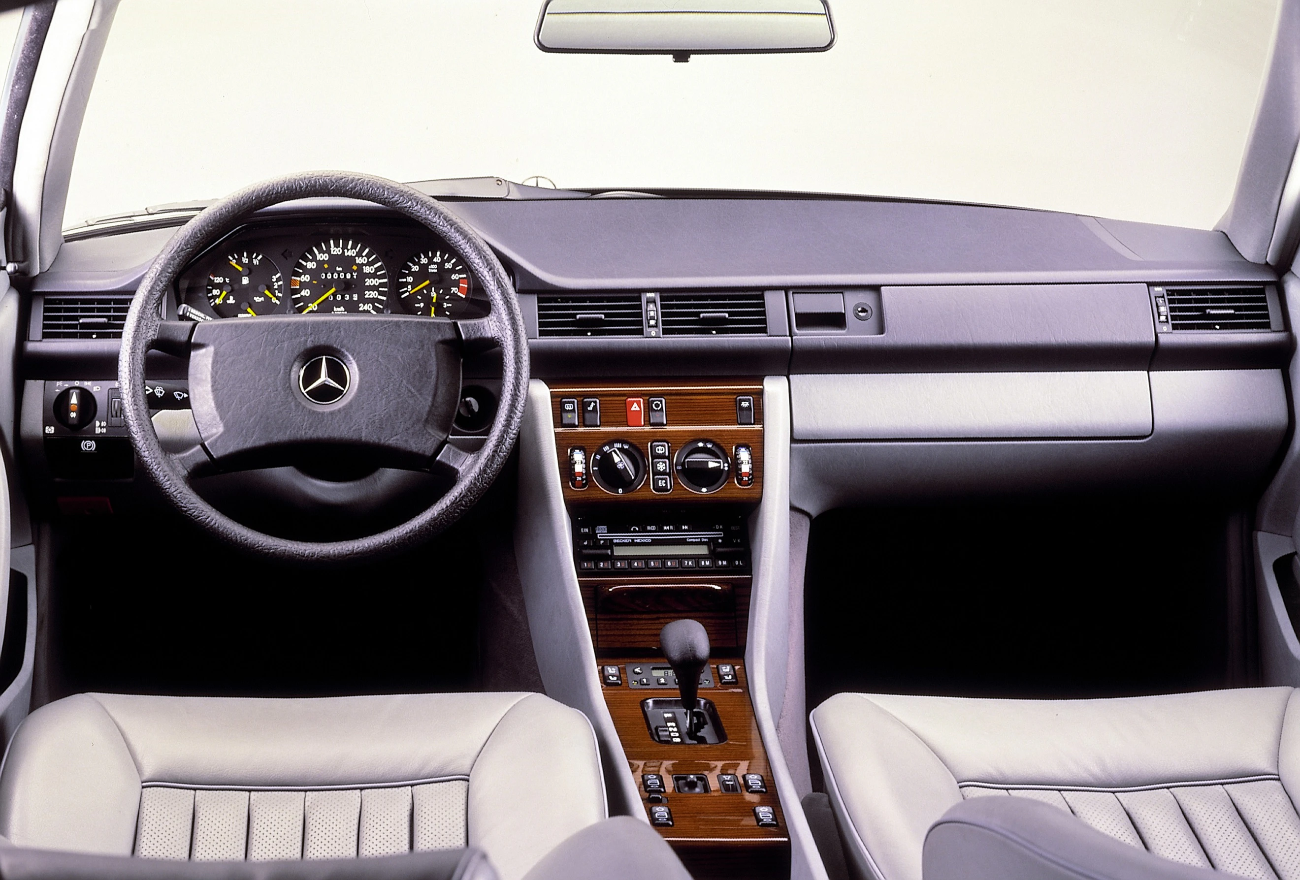 Innenraum eines klassischen Mercedes mit grauen Sitzen, Holzverkleidung und Mittelkonsole. Sicht auf Lenkrad, Armaturenbrett und Schalthebel.