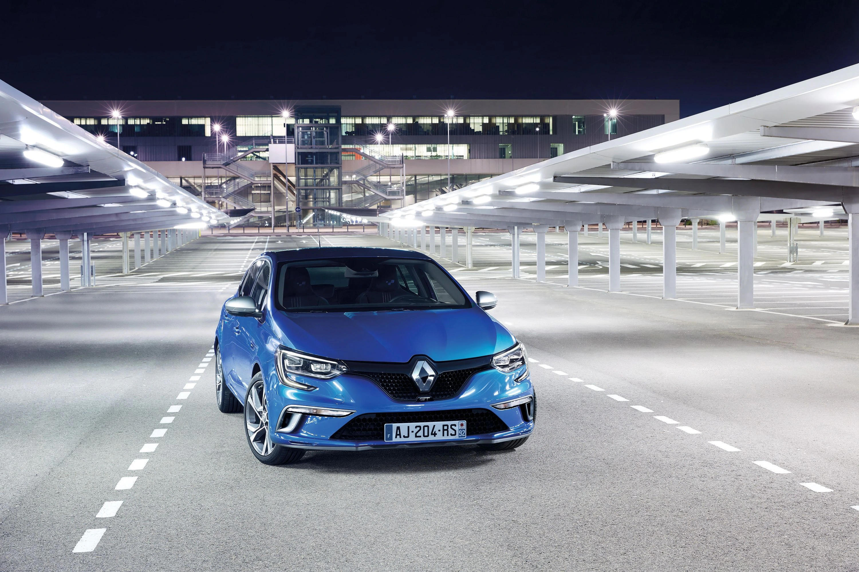 Blauer Renault Megane nachts auf beleuchtetem Parkdeck, moderne Gebäude im Hintergrund.