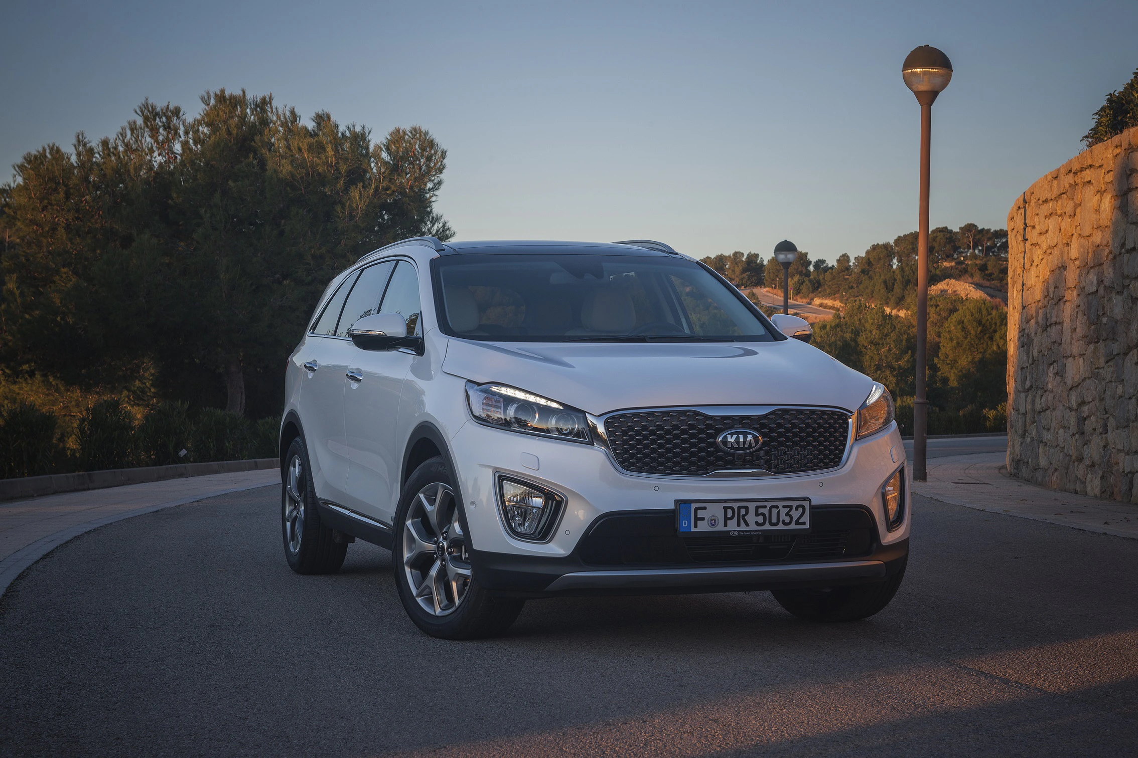 Weißer SUV steht auf einer Straße bei Sonnenuntergang, umgeben von Bäumen und einer Steinmauer. Fahrzeugfront im Mittelpunkt.
