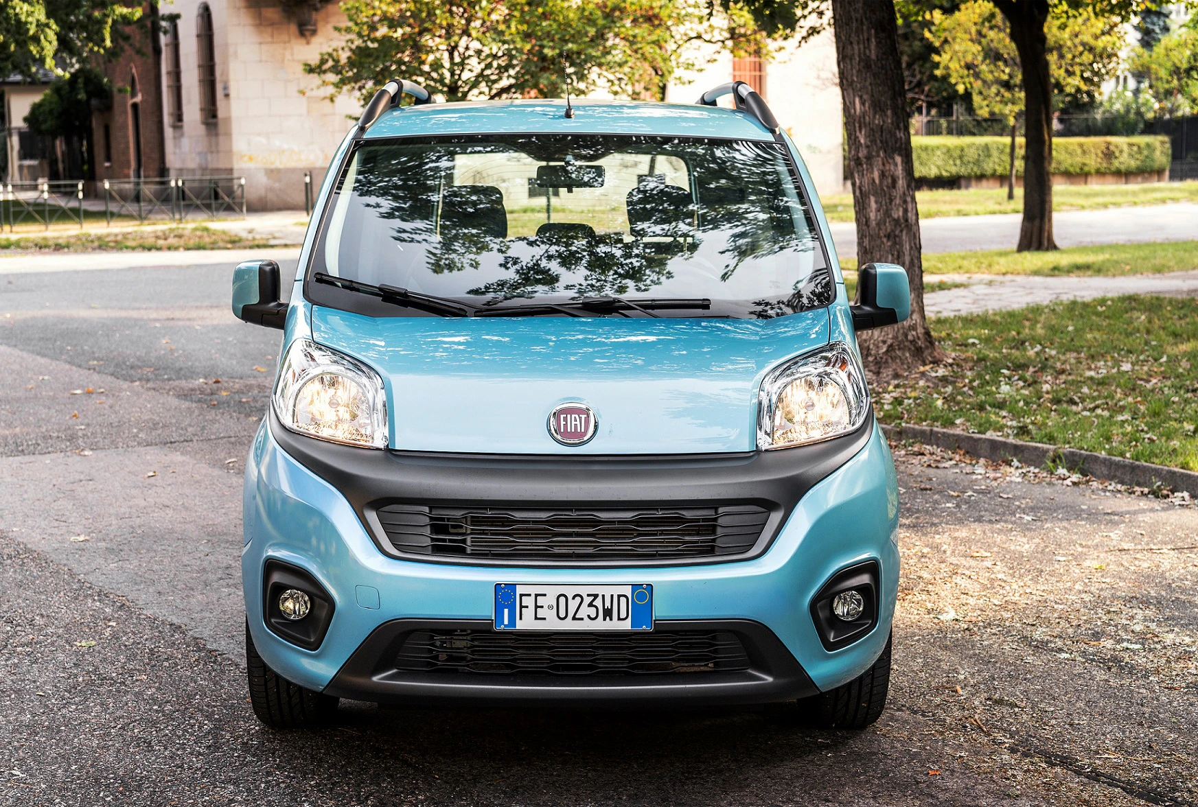 Blauer Fiat Qubo auf einer Straße im Freien, umgeben von Bäumen und Gehwegen. Frontansicht mit italienischem Kennzeichen.