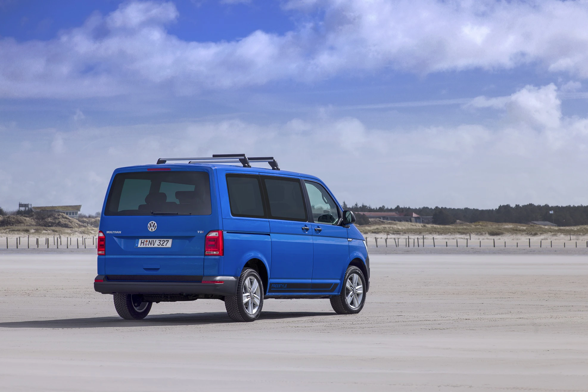 Blauer VW-Bus steht am Sandstrand unter bewölktem Himmel. Im Hintergrund sind Dünen und eine Ferienhaussiedlung zu sehen.