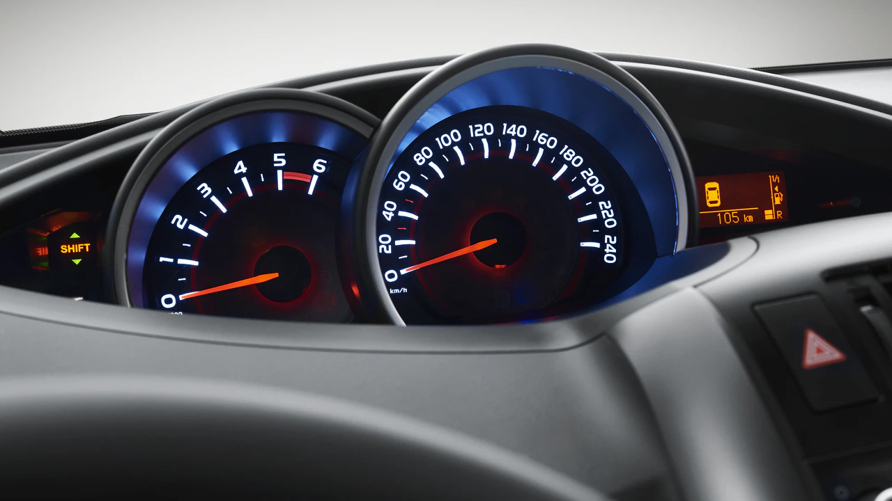 Nahaufnahme eines Autotachos mit Tachometer bei 0 km/h und Drehzahlmesser, beide von blauer Beleuchtung umgeben. Rechts ein kleines Display.
