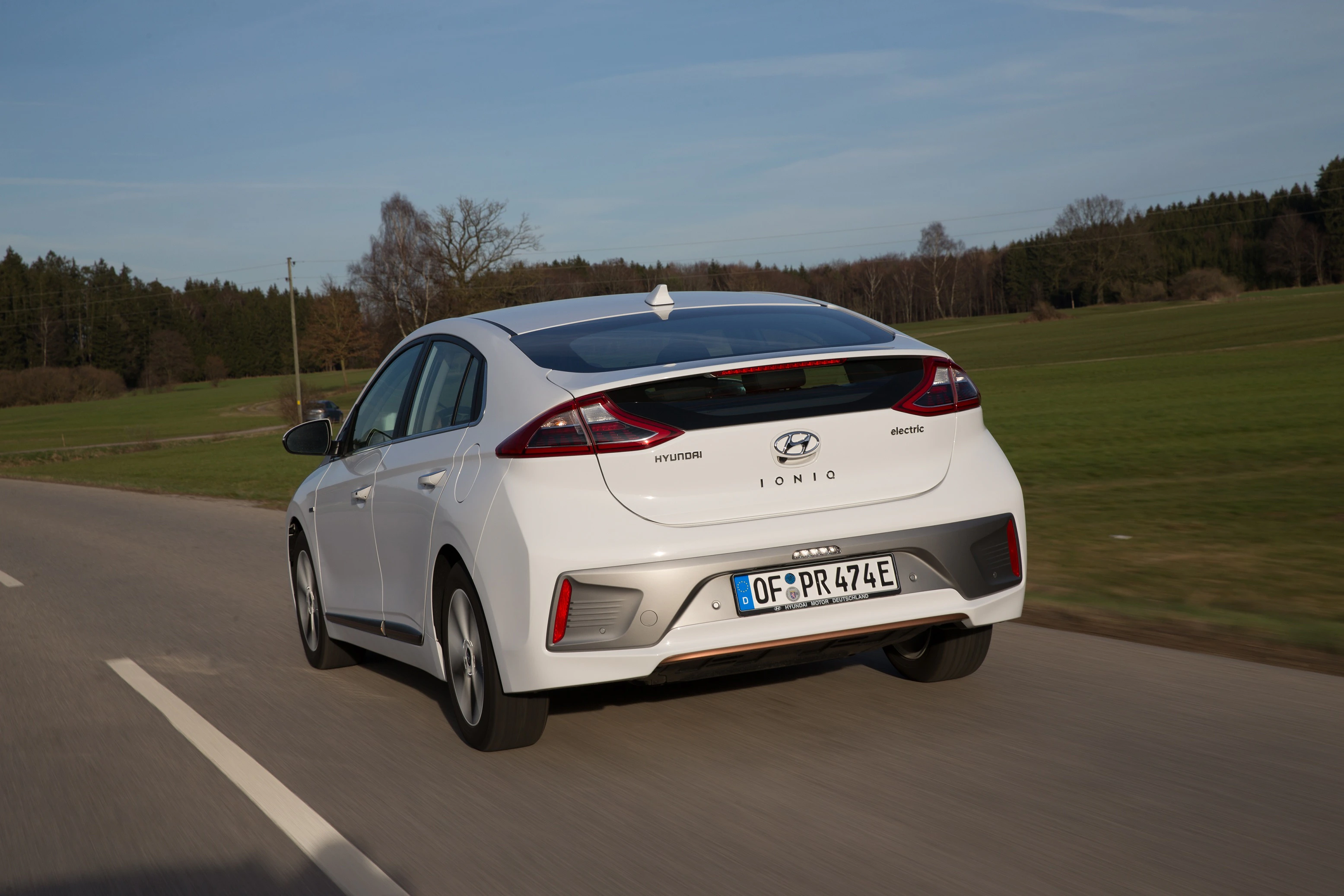 Weißer Hyundai Ioniq Hybrid fährt auf Landstraße durch grüne Landschaft, Bäume im Hintergrund.