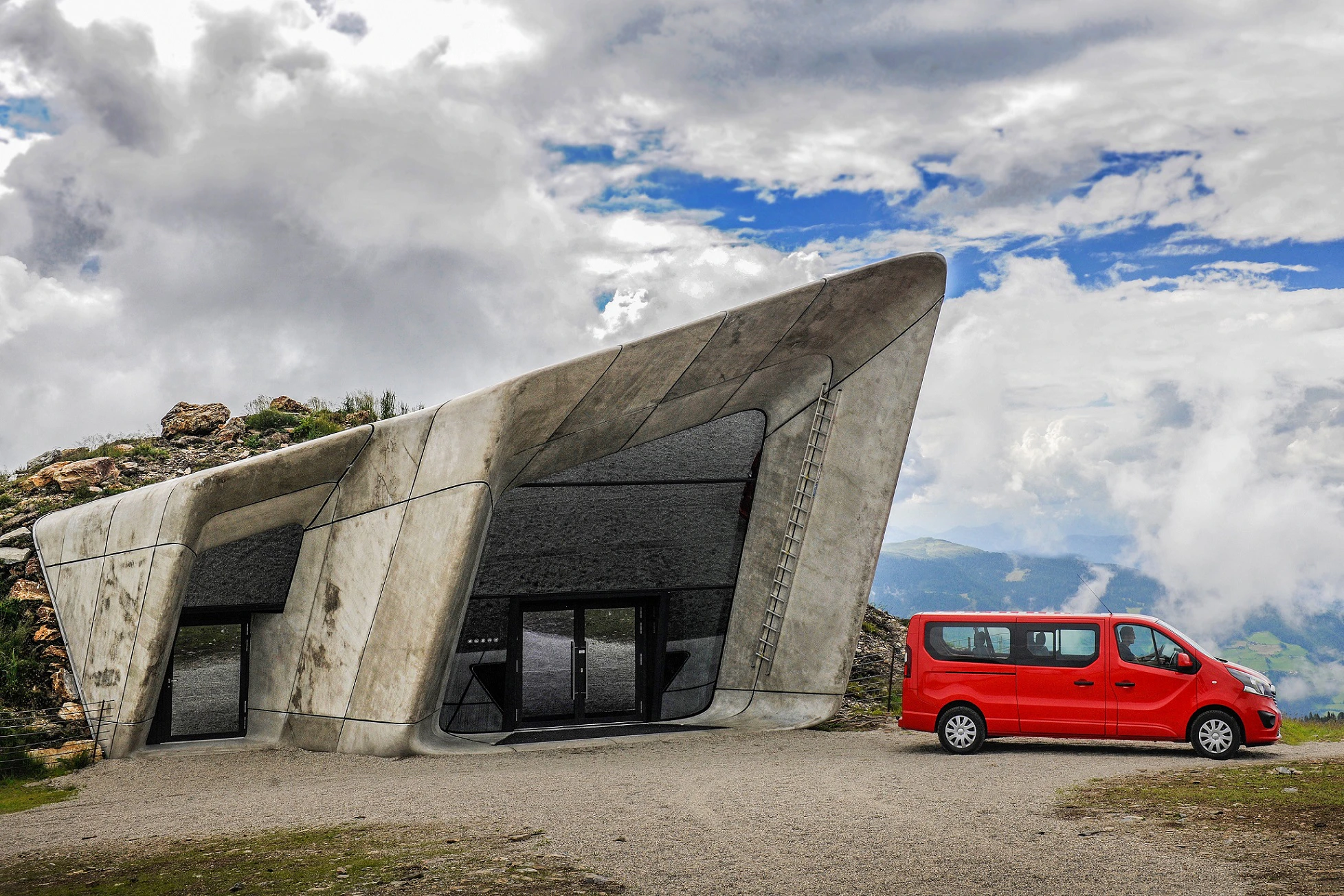 Modernes Gebäude mit schrägen Betonwänden und großen Fenstern, daneben ein roter Van, umgeben von Wolken und Bergen.