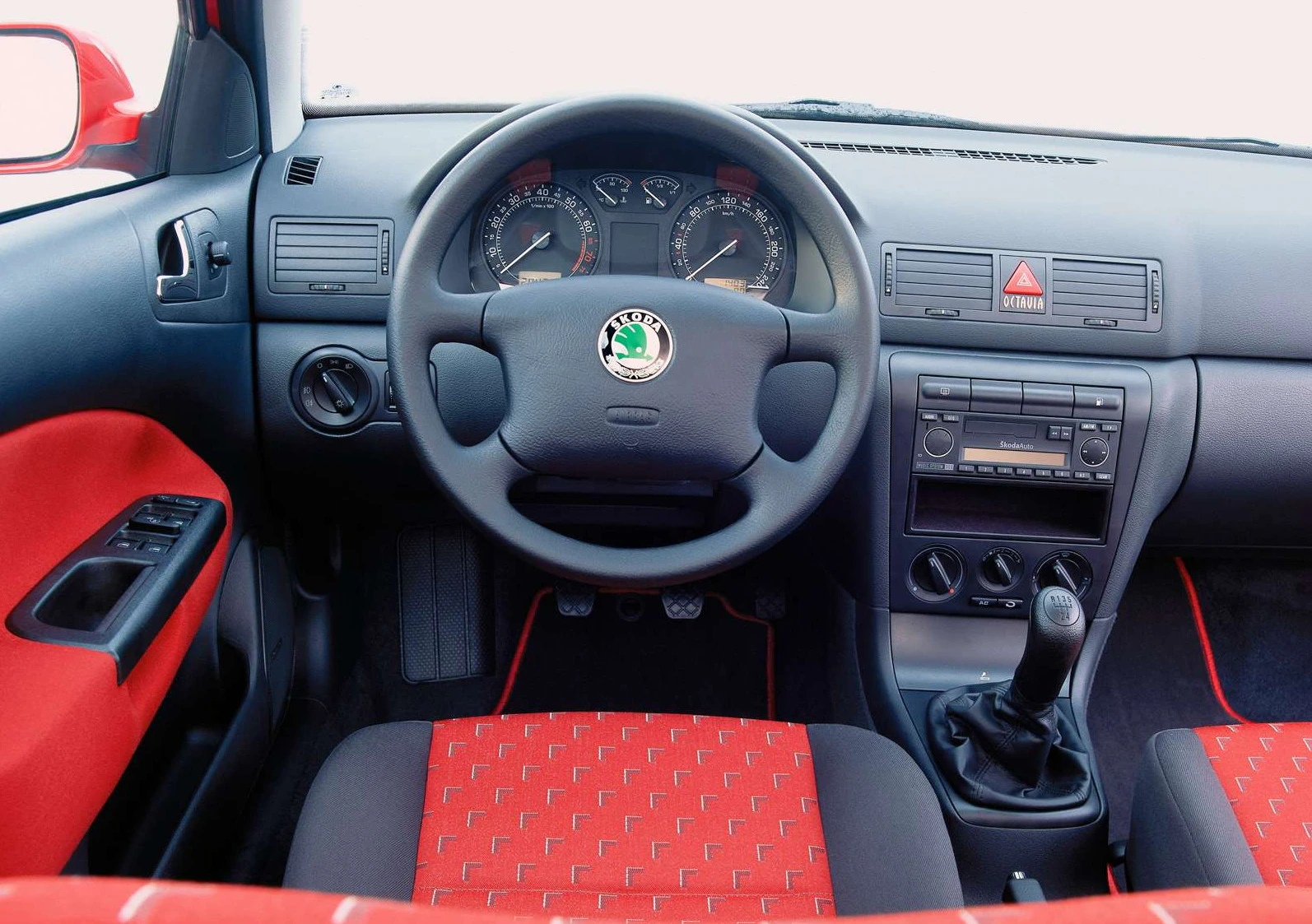 Innenraum eines Autos mit schwarzem Armaturenbrett, Lenkrad und roten Sitzen. Zeigt Tacho, Lüftung, Radio und manuelle Gangschaltung.