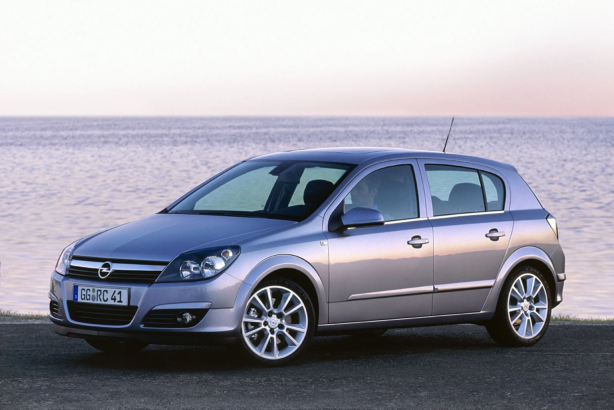Silberner Opel Astra parkt vor einem ruhigen See bei Sonnenuntergang. Der Himmel spiegelt sich im Wasser wider.
