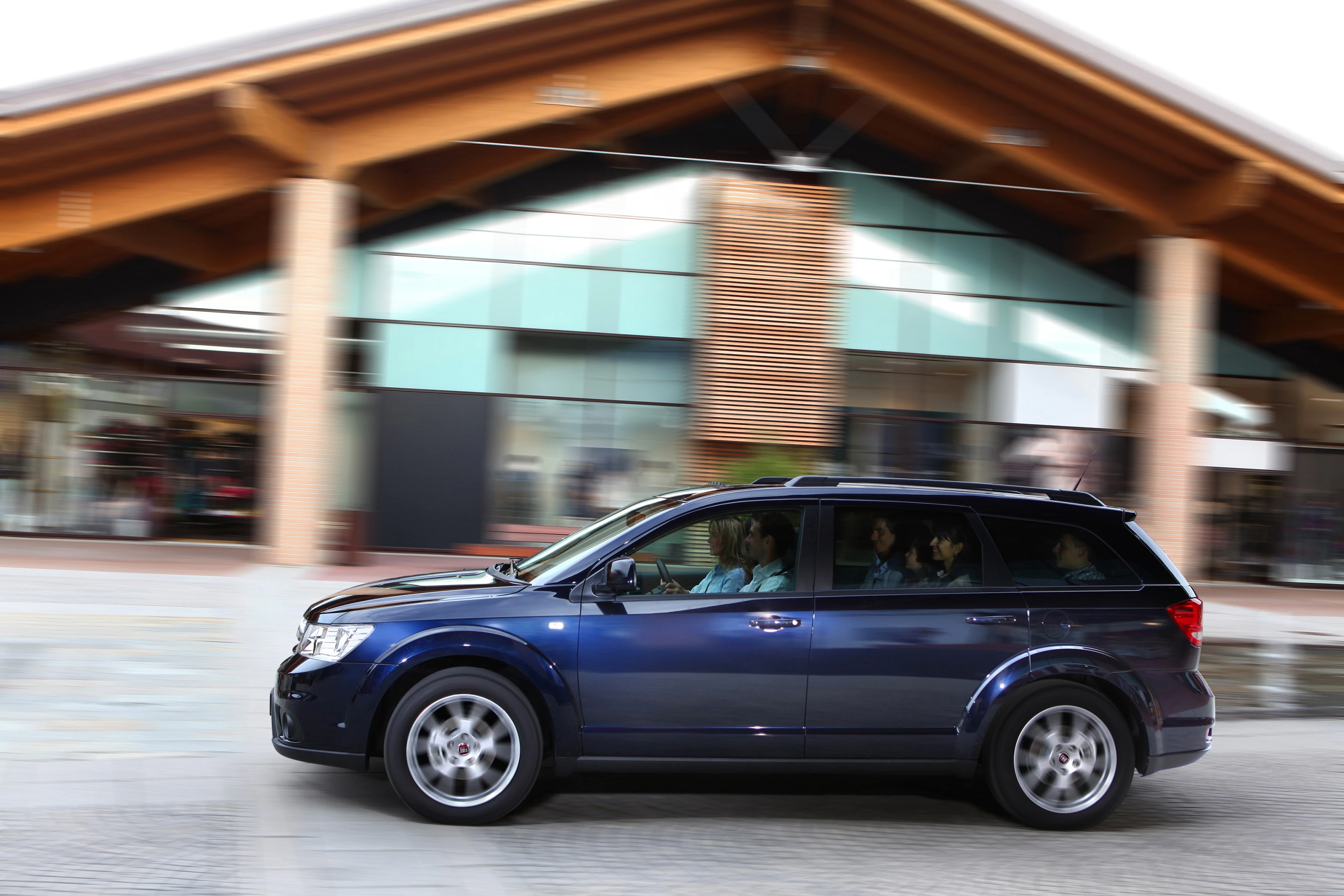 Blauer SUV fährt schnell an modernen Gebäuden vorbei. Passagiere sind im Auto sichtbar.