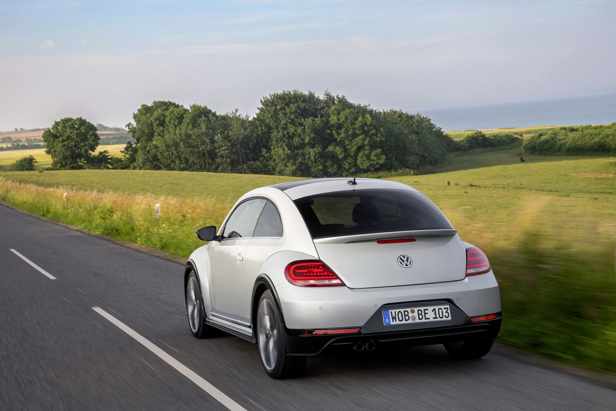 Silberner Volkswagen Beetle fährt auf einer Landstraße durch grüne Landschaft, Bäume und Felder im Hintergrund.