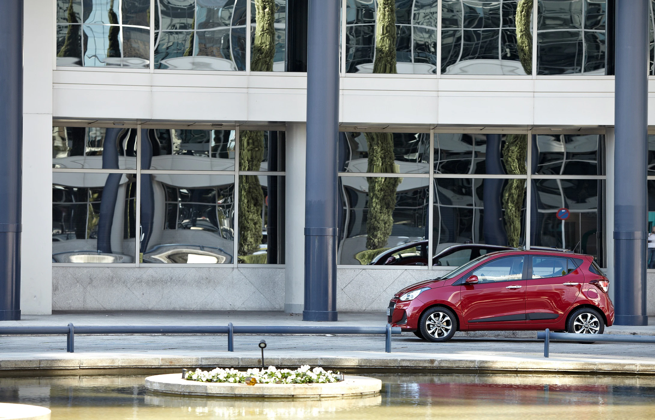 Rotes Auto vor einem modernen Gebäude mit Glasfassade, die Bäume und Säulen spiegelt, in urbaner Umgebung.