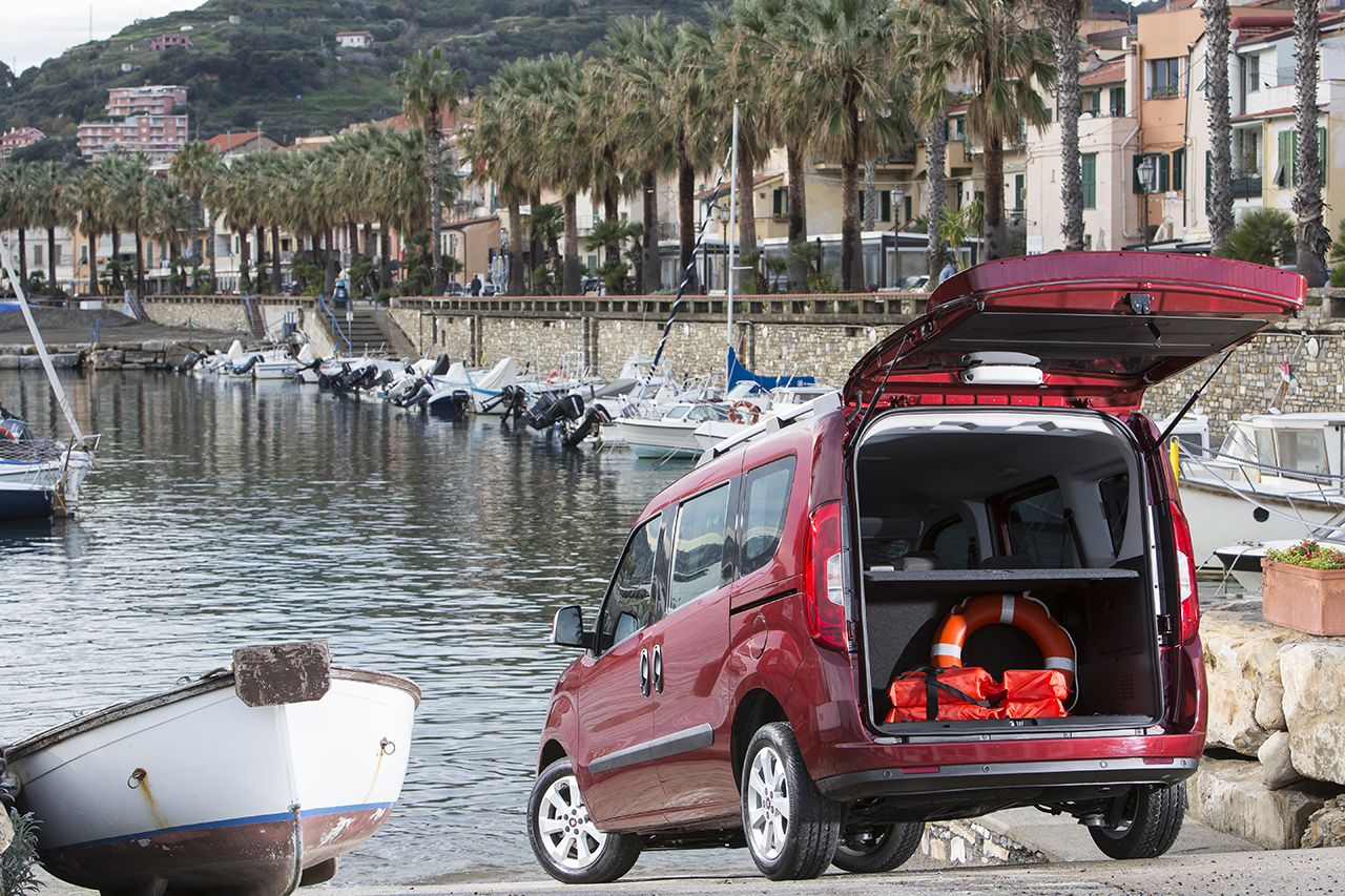 Rotes Auto mit offener Heckklappe am Wasser. Boote auf dem Wasser, Palmen und Häuser im Hintergrund. Rettungsring im Kofferraum.