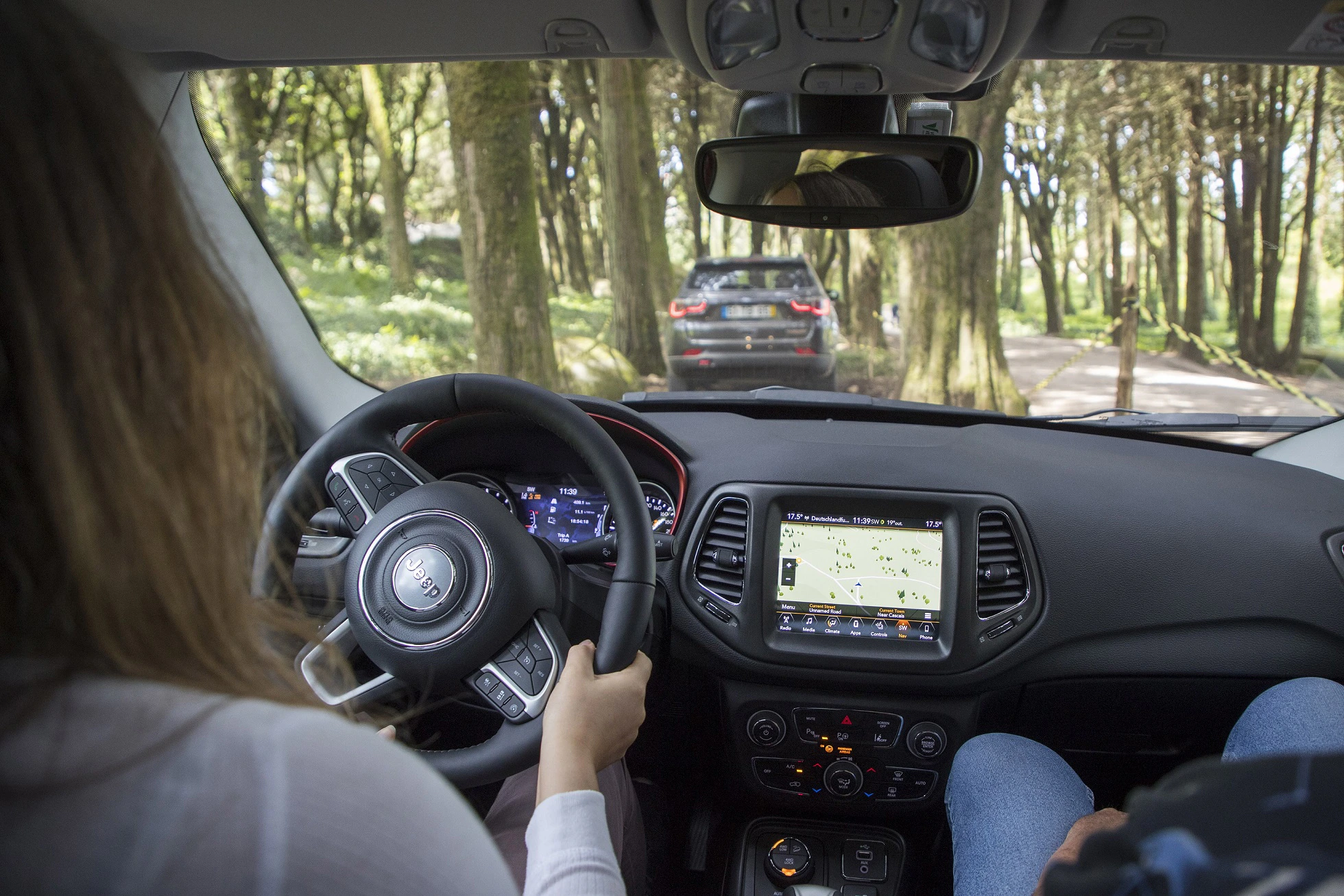 Blick aus dem Auto auf gewundene Waldstraße. Fahrerin am Lenkrad, Navi auf Display, Auto voraus im Grünen.