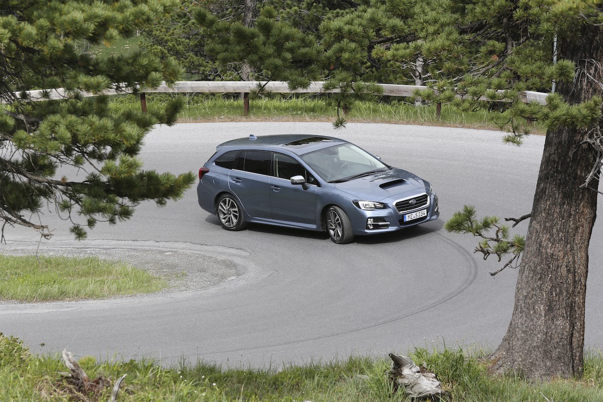 Blauer Kombi fährt durch eine enge Kurve auf einer Bergstraße, umgeben von Bäumen und Wiesen.