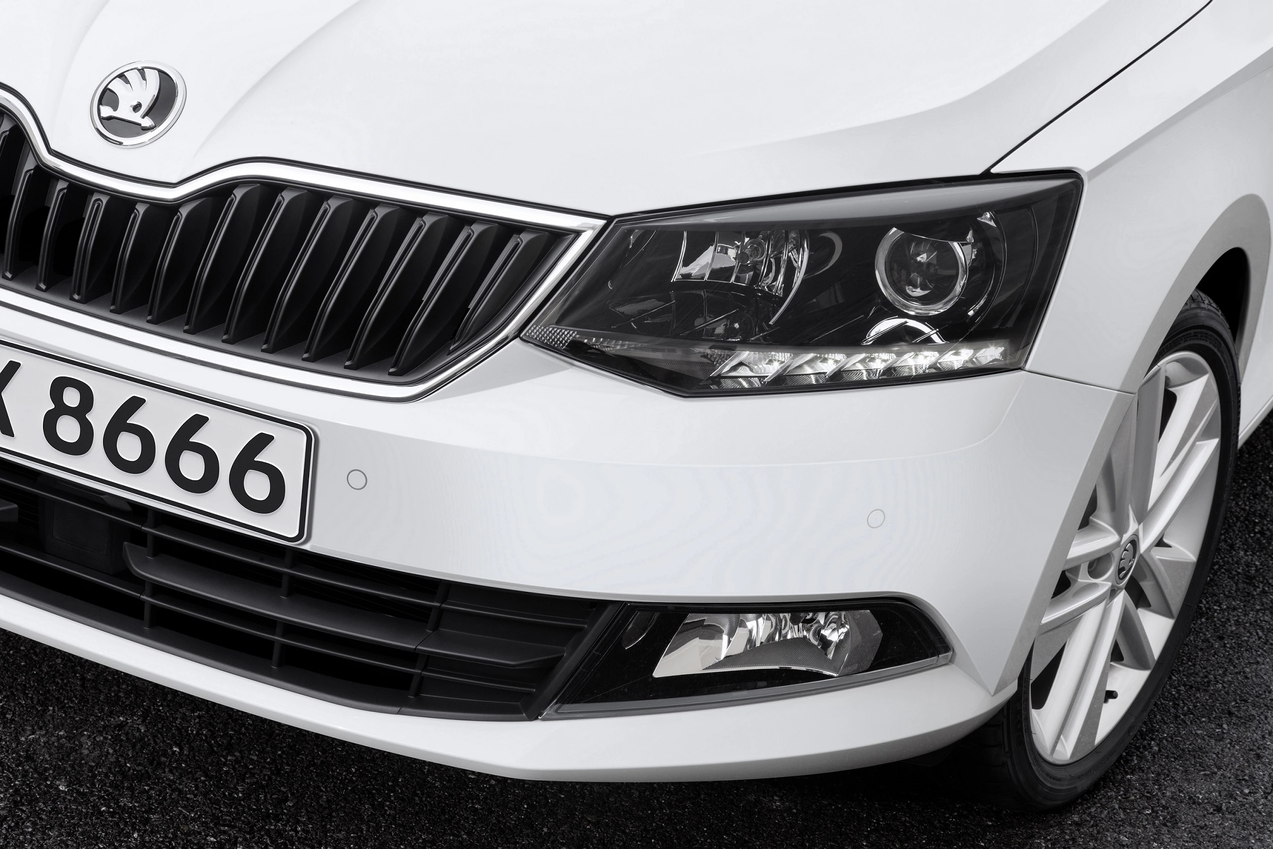 Nahaufnahme eines weißen Škoda-Autos mit modernem Scheinwerferdesign und Nummernschild mit der Nummer "X 8666" auf Asphalt.