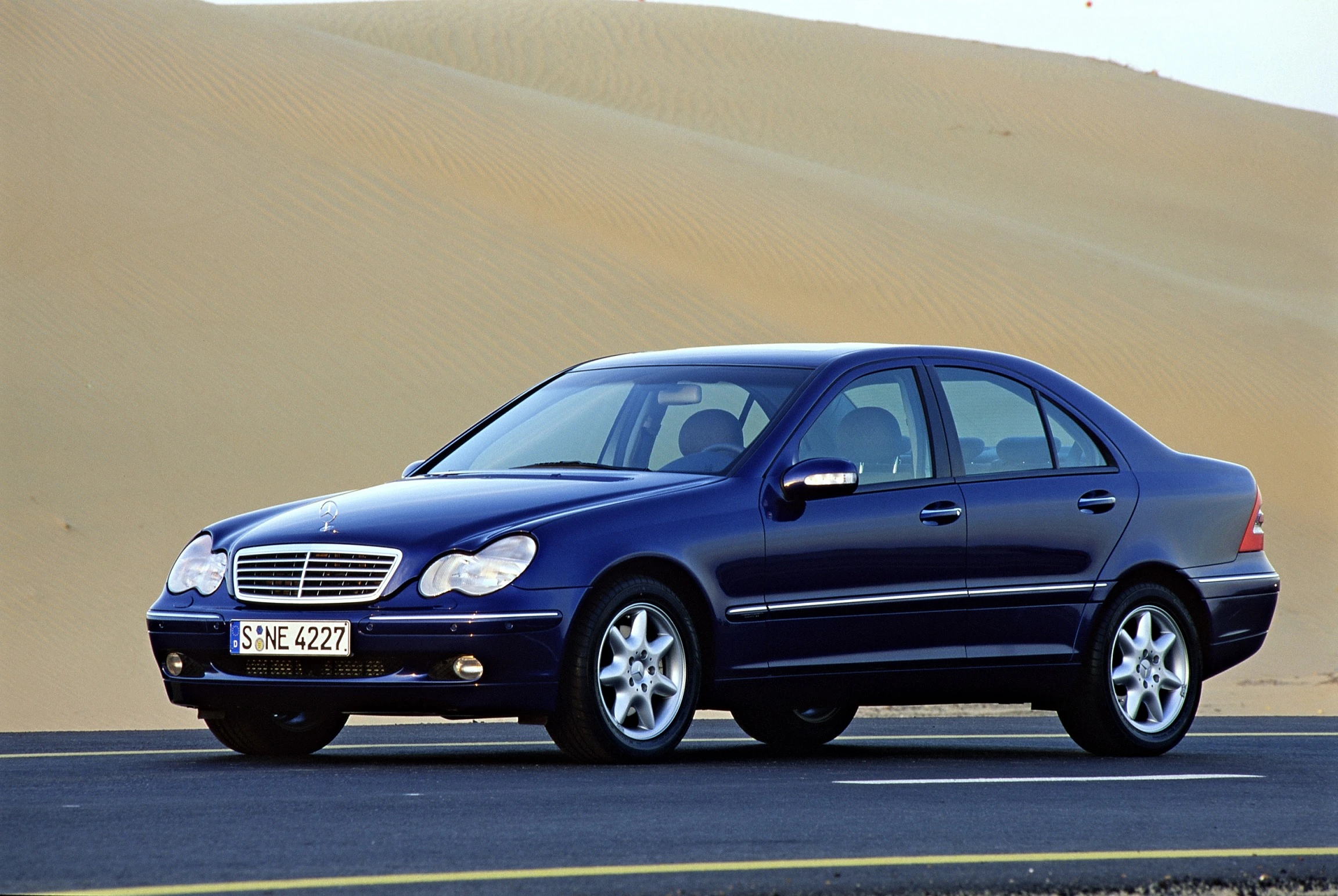 Blauer Mercedes-Benz C-Klasse Limousine auf einer asphaltierten Straße, umgeben von Sanddünen in einer Wüstenlandschaft.