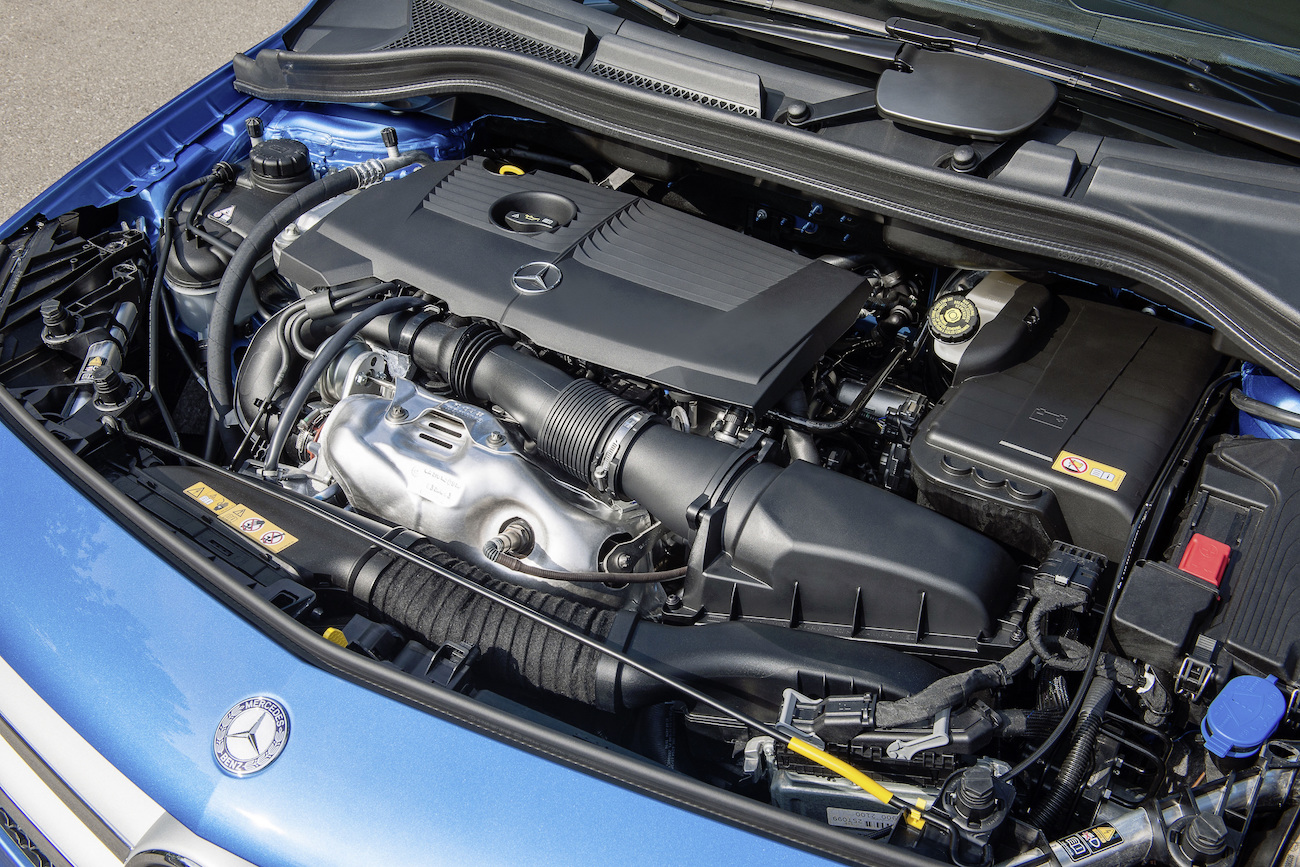 Motorraum eines blauen Autos mit geöffnetem Deckel, sichtbar ist ein moderner Motor mit dem Mercedes-Logo und verschiedenen Autoteilen.