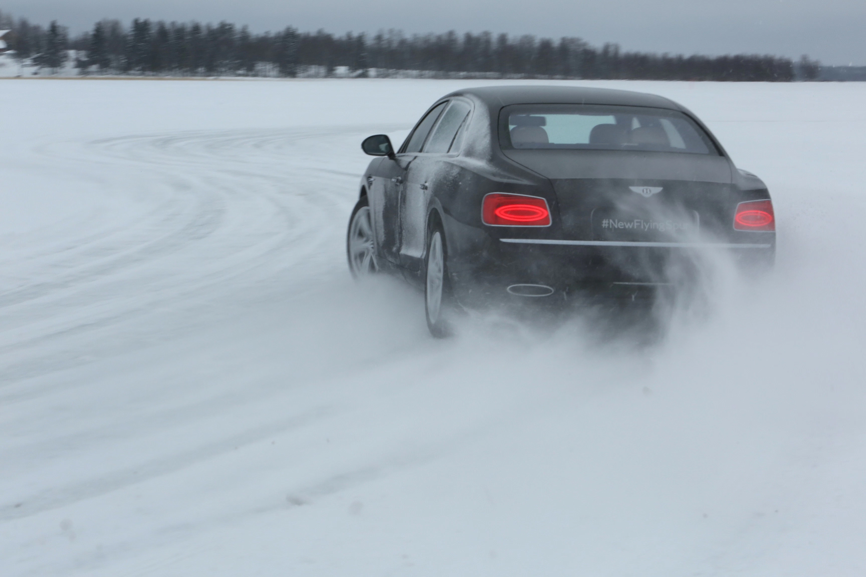 Ein schwarzes Auto fährt auf einer schneebedeckten Strecke, wirbelt Schnee auf. Im Hintergrund sind Bäume zu sehen.