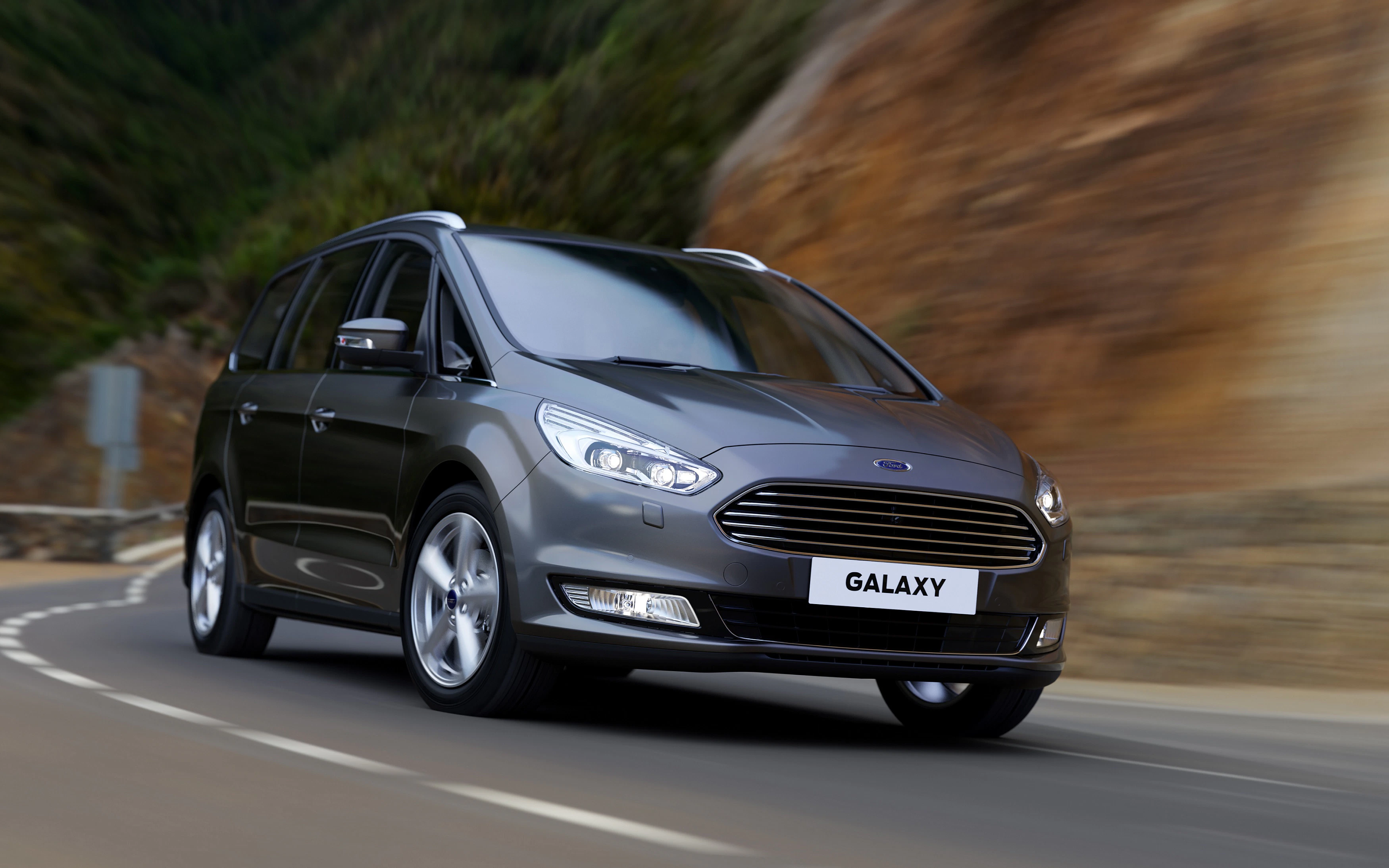 Grauer Ford Galaxy fährt auf kurviger Bergstraße. Dynamische Perspektive. Klarer Himmel und felsige Landschaft im Hintergrund.