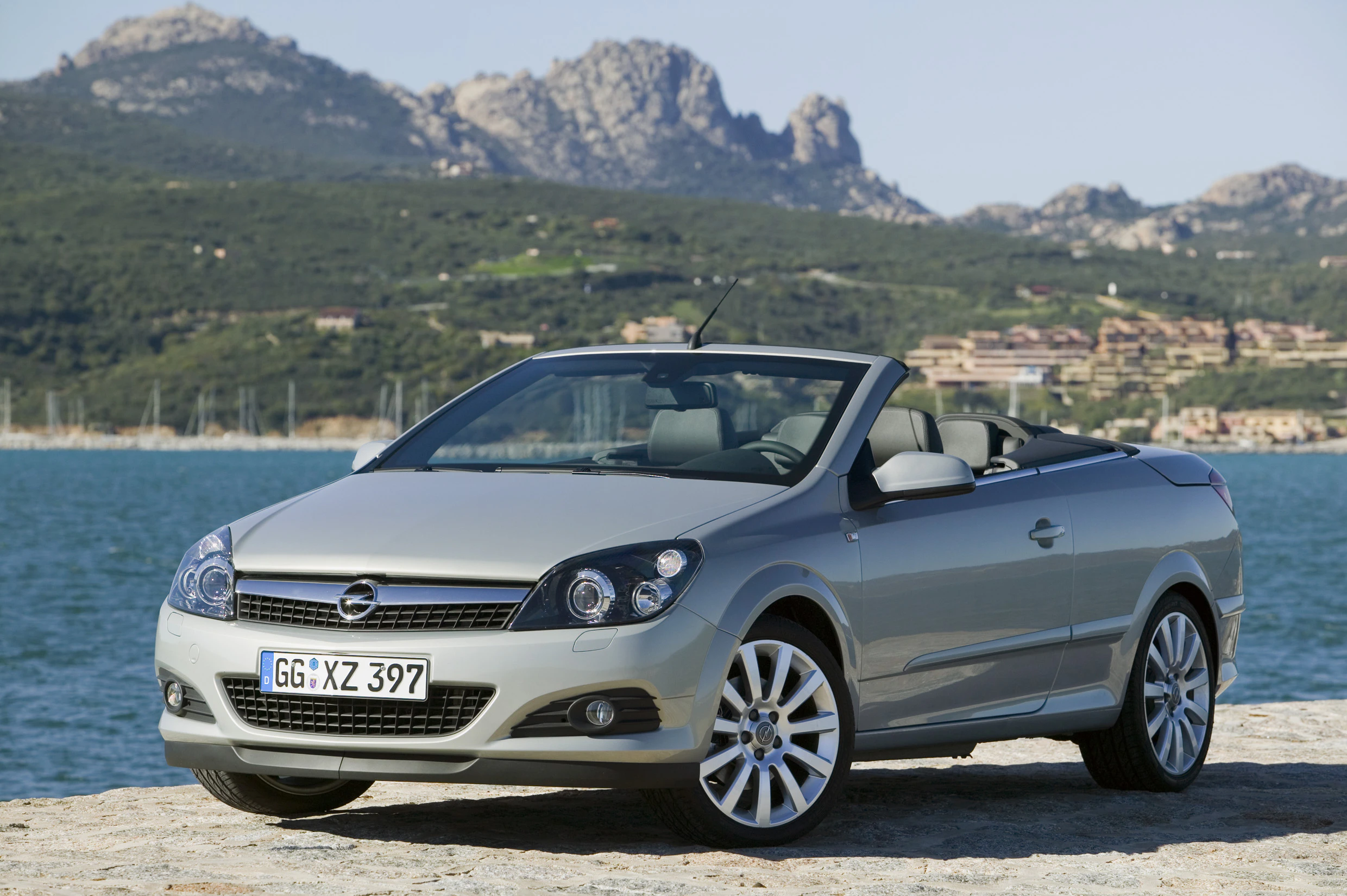 Silbernes Cabrio geparkt vor einer malerischen Küstenlandschaft mit Bergen im Hintergrund. Klarer Himmel und ruhiges Wasser.