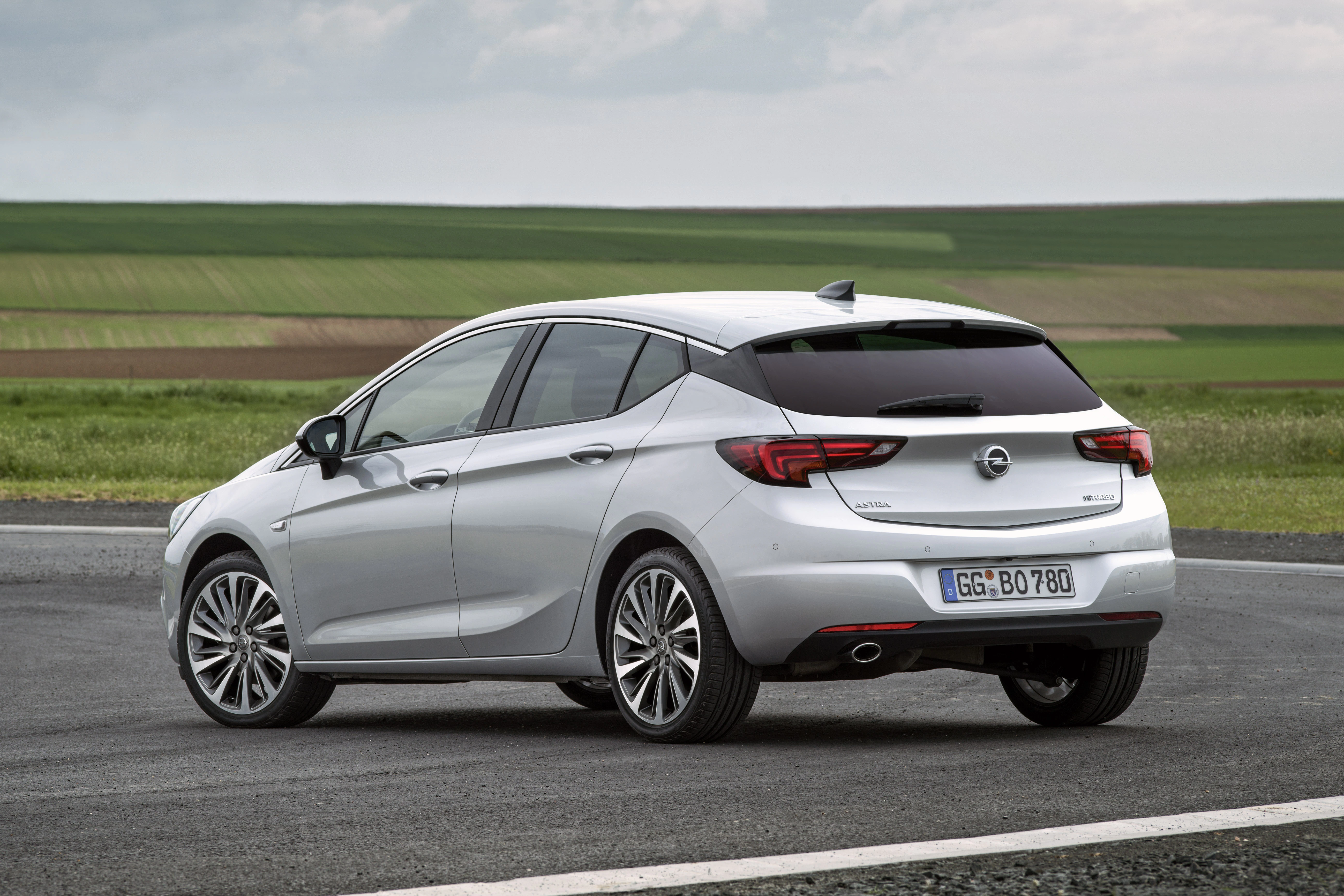 Silberner Opel Astra parkt auf einer asphaltierten Fläche vor einer grünen, ländlichen Landschaft unter bewölktem Himmel.