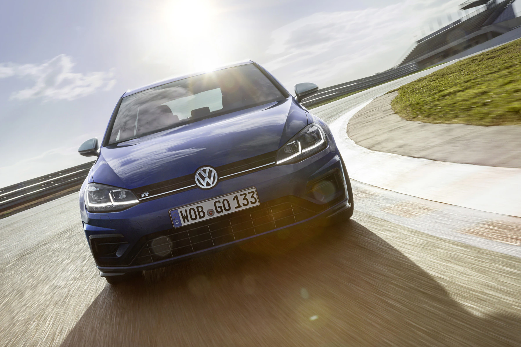 Blauer Volkswagen fährt dynamisch auf einer Rennstrecke bei Sonnenschein. Frontansicht, mit deutlichem Markenzeichen.