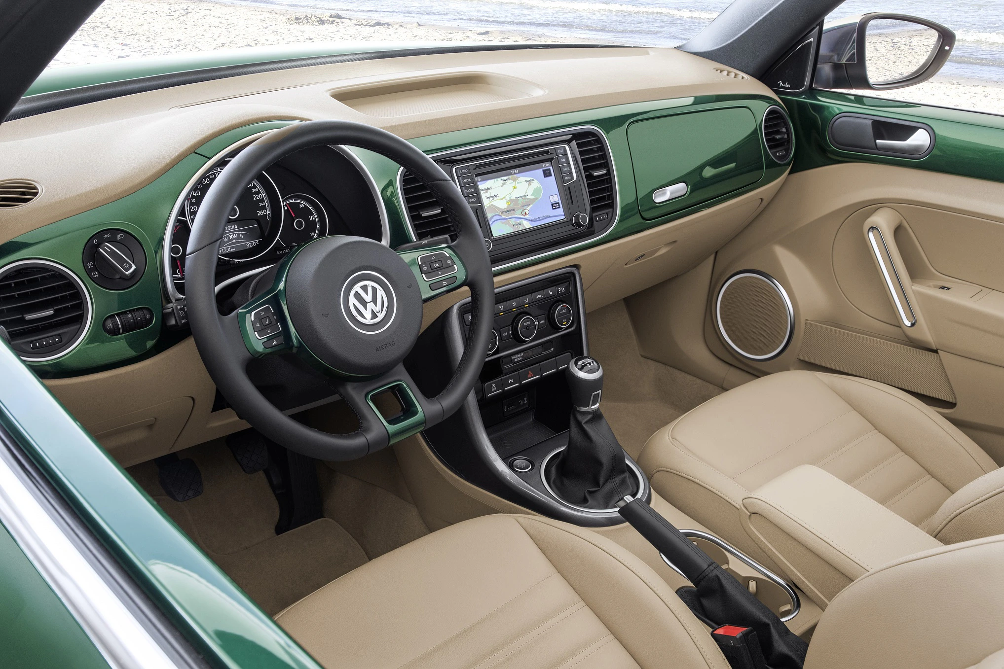 Innenraum eines grünen Volkswagen mit braunen Ledersitzen, Lenkrad, Schaltknüppel und Touchscreen. Blick vom Fahrersitz aufs Meer.