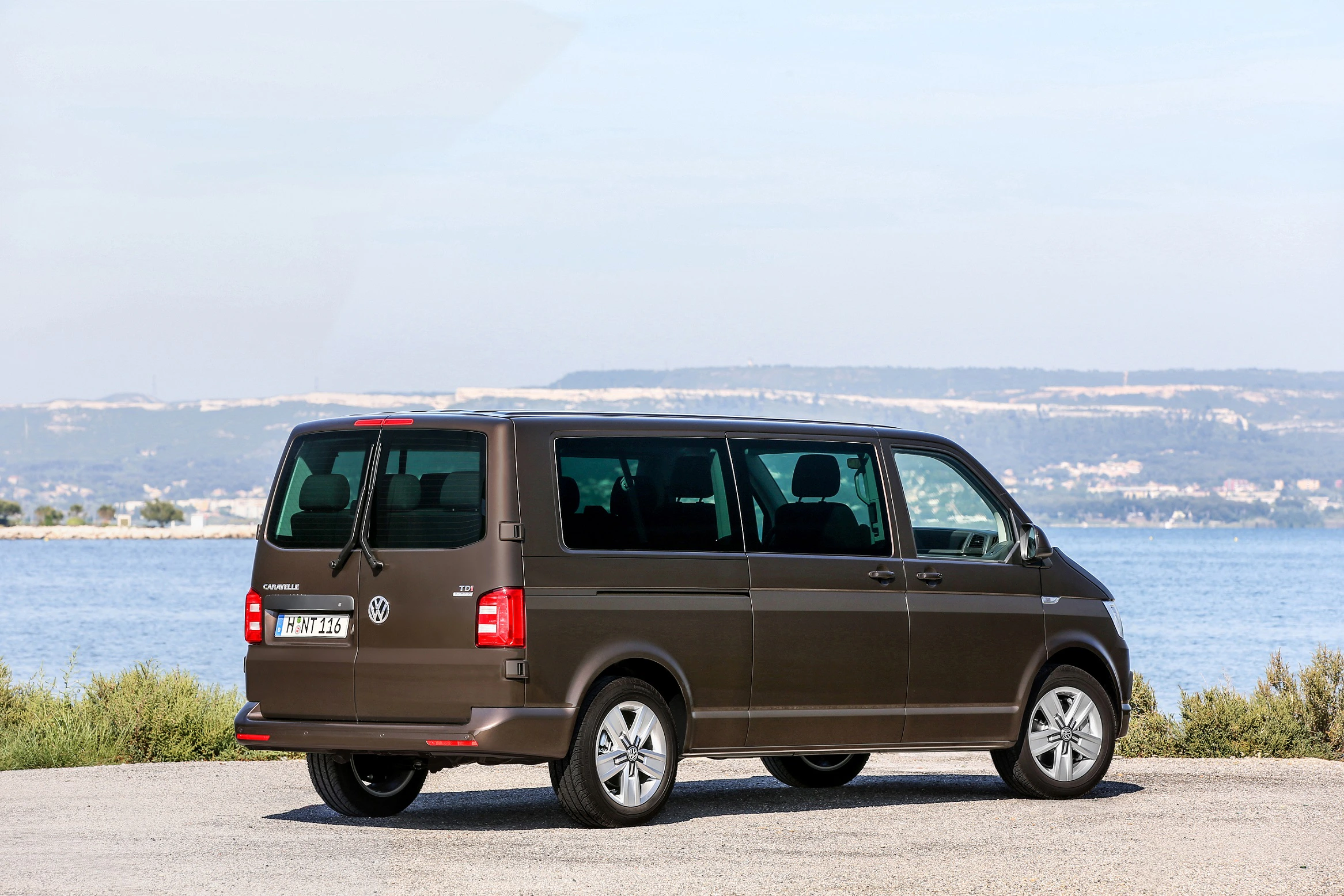 Brauner Minivan, parkend vor einer Küstenlandschaft mit blauem Meer und Hügeln im Hintergrund. Klarer Himmel und Sonnenschein.